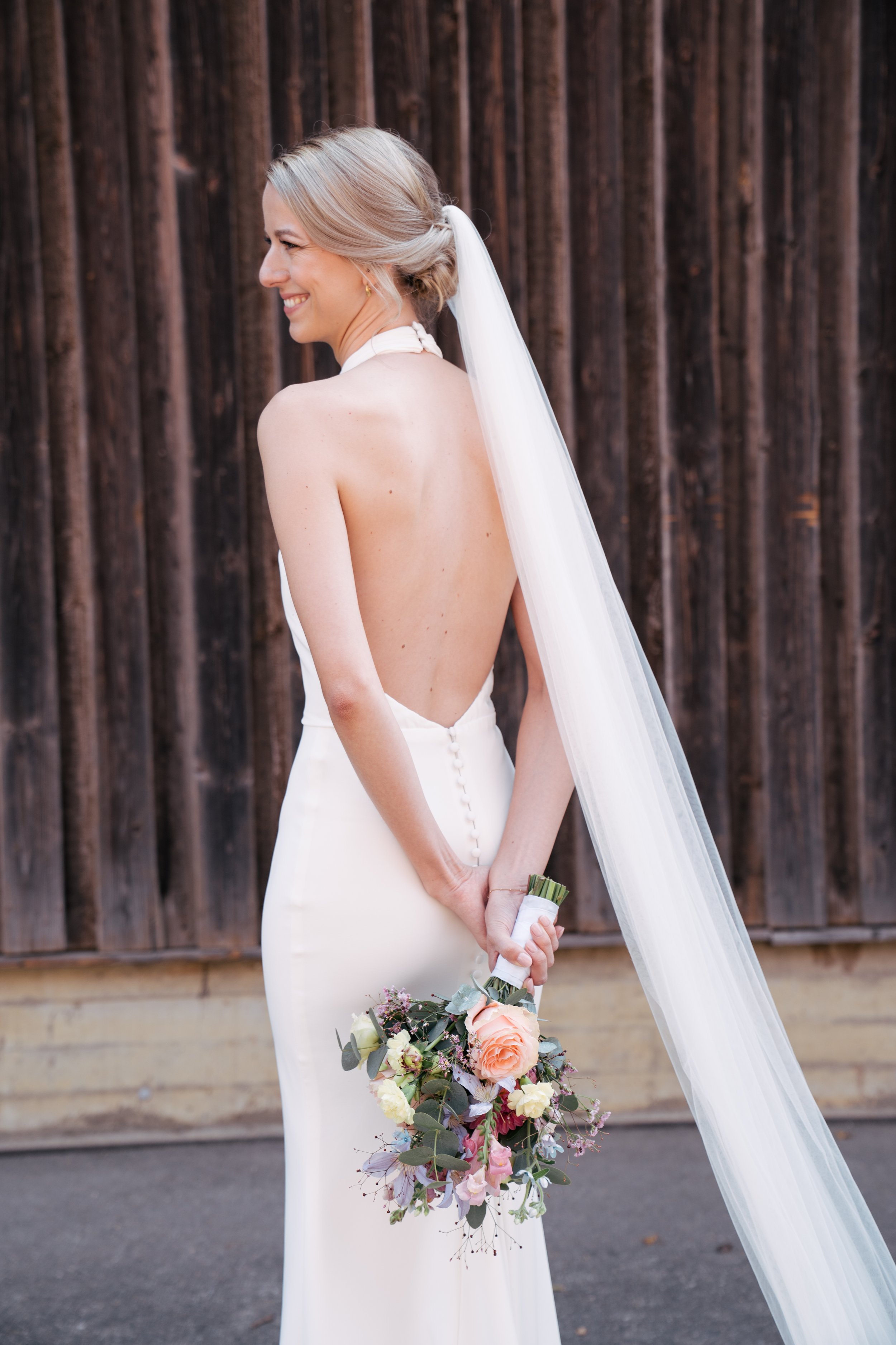 Eine lächelnde Braut in einem weißen Kleid mit offenem Rücken hält einen Blumenstrauß, während sie vor einer Holzverschalung steht.