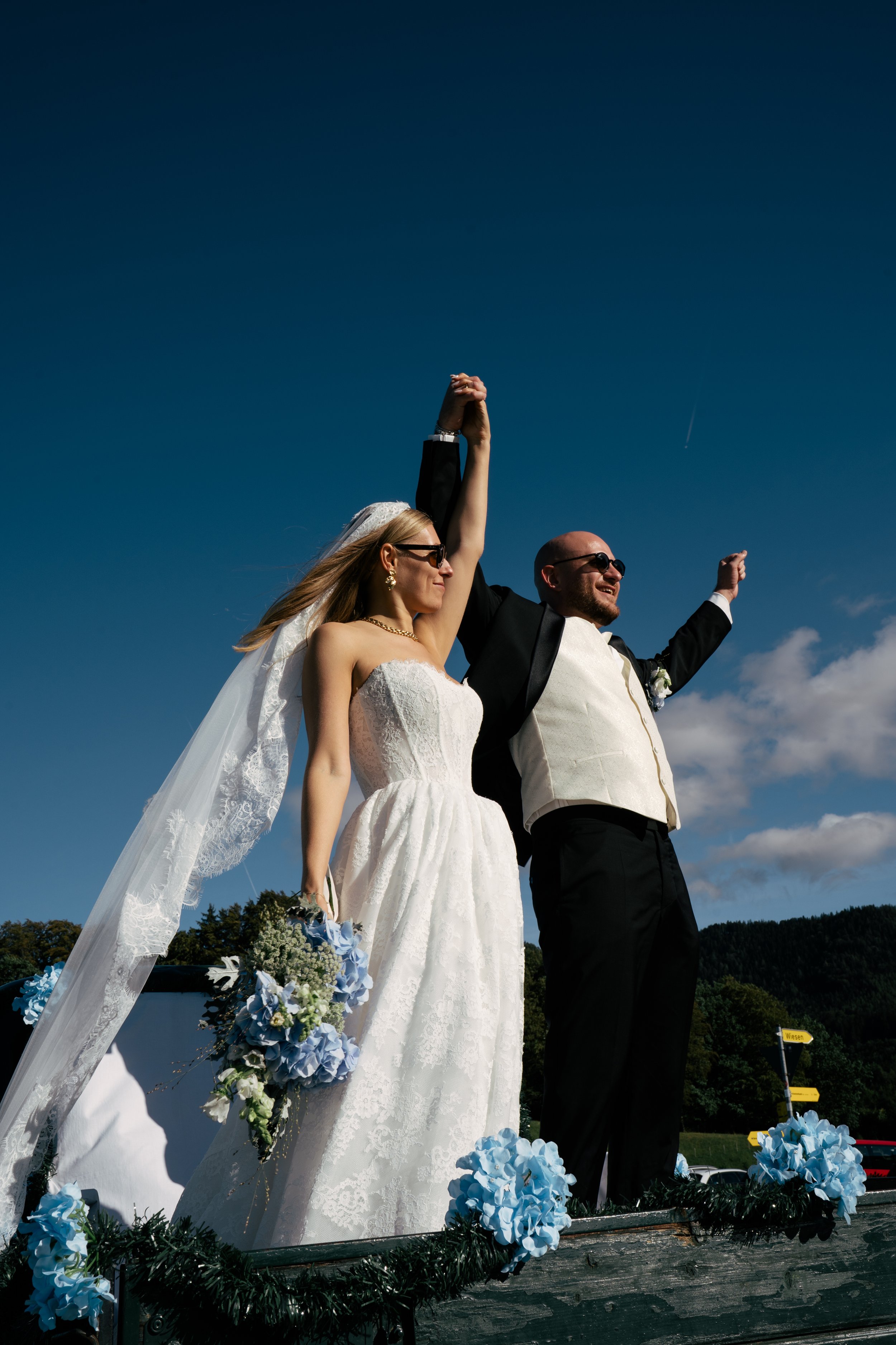 Ein frisch vermähltes Paar, ein Braut und ein Bräutigam, steht auf einer festlich geschmückten Plattform im Freien, beide mit Sonnenbrillen, die Arme erhoben in Siegerpose. Die Braut trägt ein weißes Hochzeitskleid mit Schleier, die Braut hält einen Blumenstrauß, der Bräutigam trägt einen schwarzen Anzug. Der Himmel ist blau mit wenigen Wolken. Im Hintergrund sind Bäume und eine Richtungsangabe "Wiesen" sichtbar.