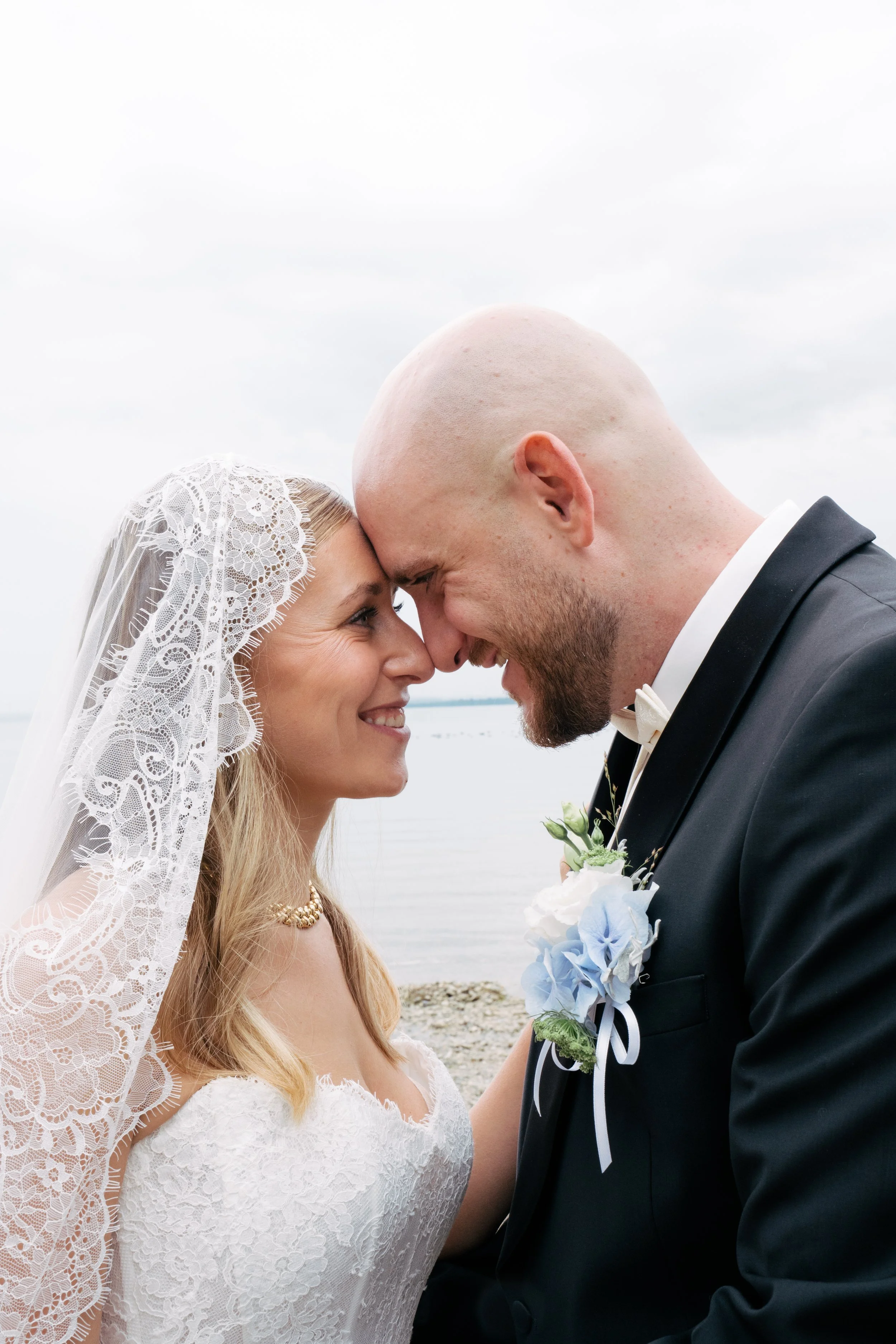 Ein brautpaar bei einer Hochzeit am Strand, die Braut trägt ein Spitzenkleid und eine Spitzenhaube, der Bräutigam trägt einen schwarzen Anzug mit Fliege, sie berühren sich mit den Stirnen und lächeln sich an.