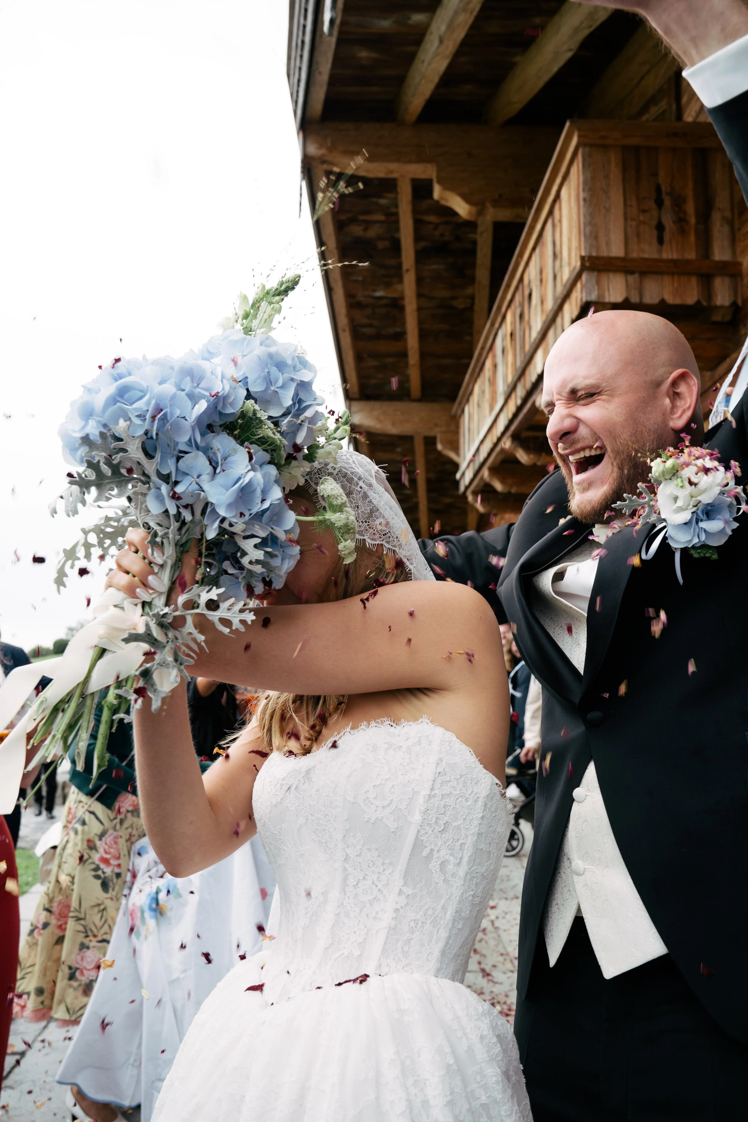 Ein Brautpaar beim Hochzeitsfest, die Frau hält einen Blumenstrauß hoch, der Mann trägt einen schwarzen Anzug mit weißer Weste und Krawatte. Es gibt Konfetti im Flug, im Hintergrund sind weitere Gäste und eine Holzkonstruktion.