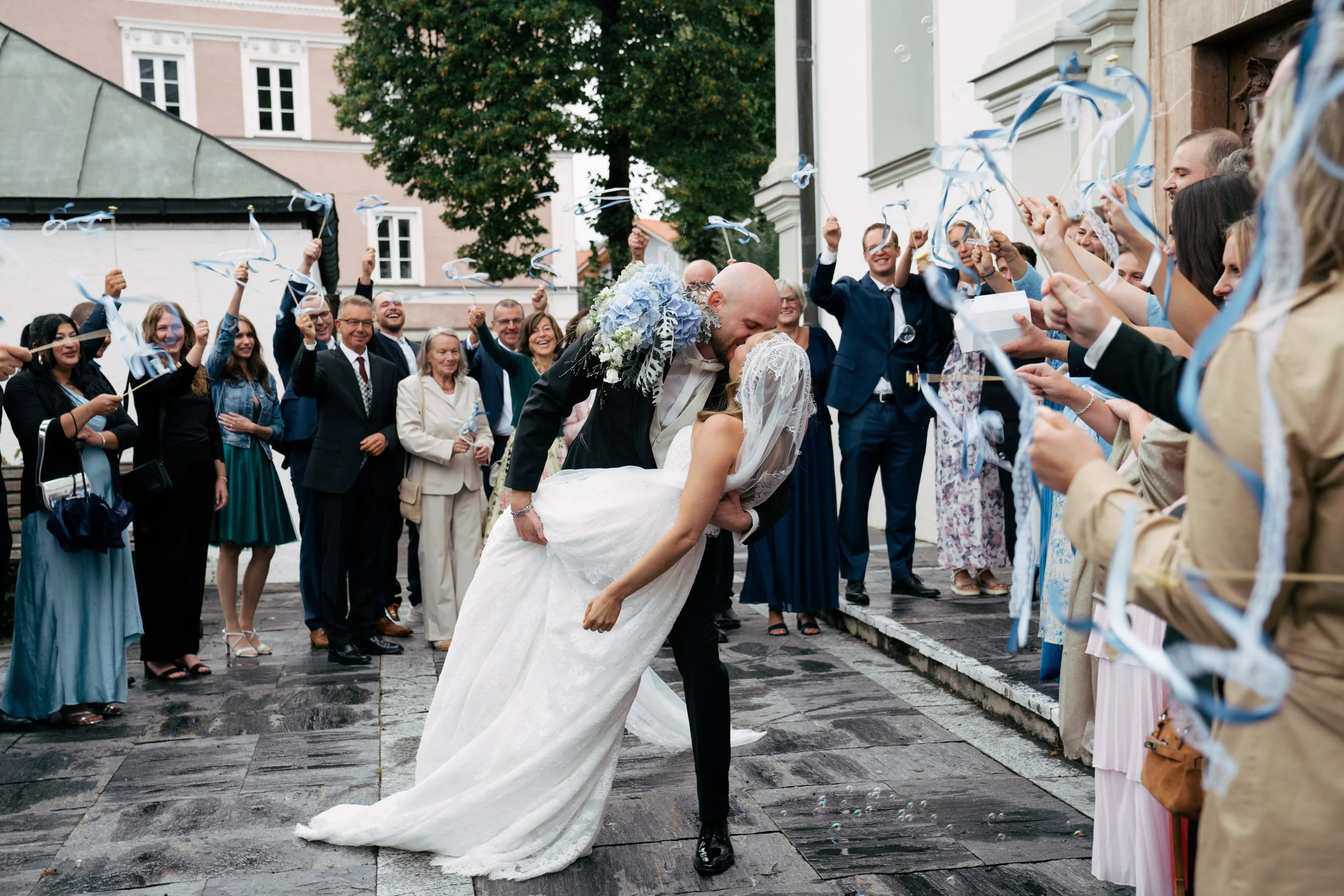 Ein Brautpaar küsst sich bei ihrer Hochzeit vor einer Gruppe von Gästen, die mit blauen und weißen Bändern feiern, vor historischen Gebäuden und einer großen Baum im Hintergrund.