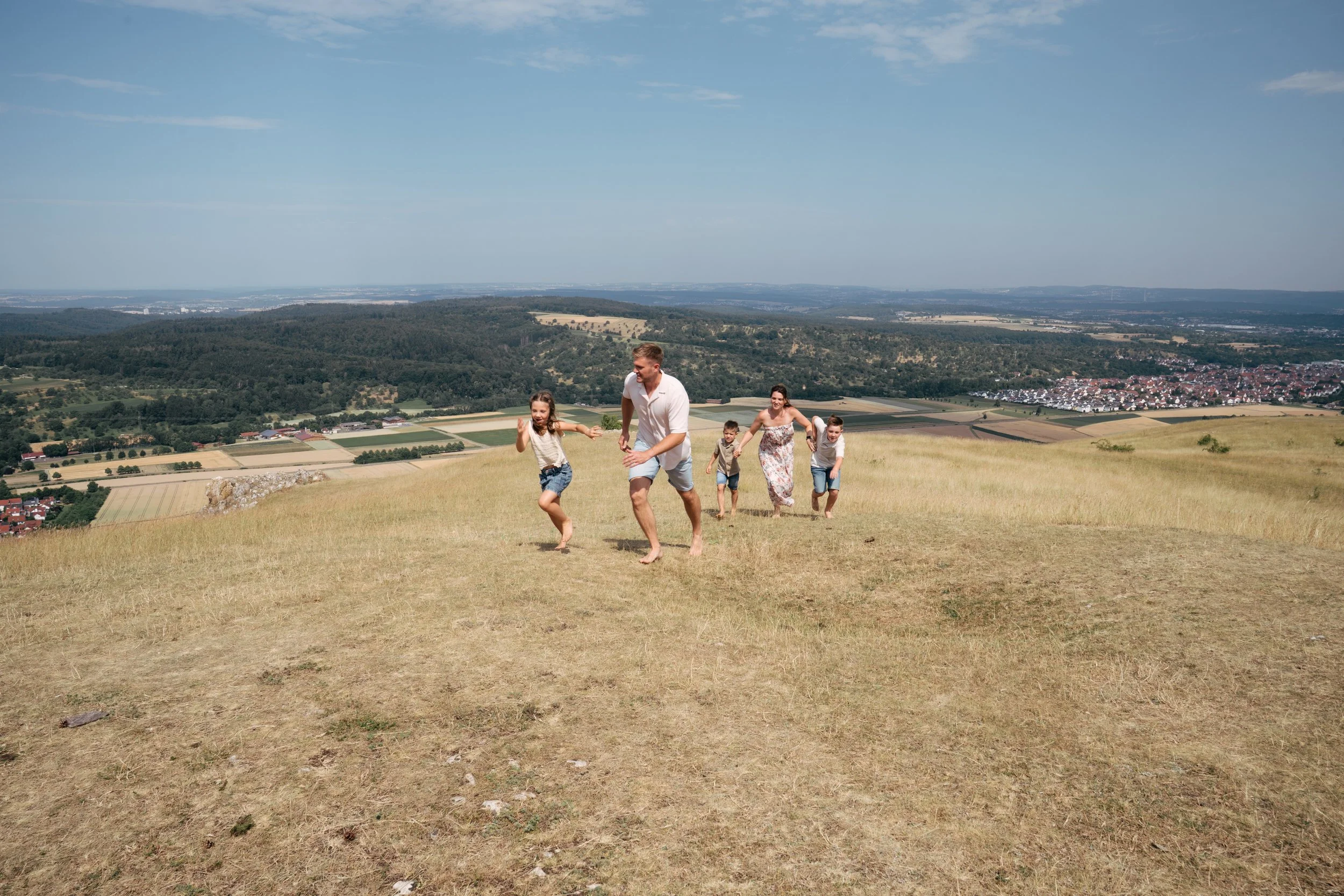 Familie beim Spielen auf einer hügeligen Wiese mit Blick auf eine Landschaft und eine Stadt im Hintergrund, bei sonnigem Wetter.