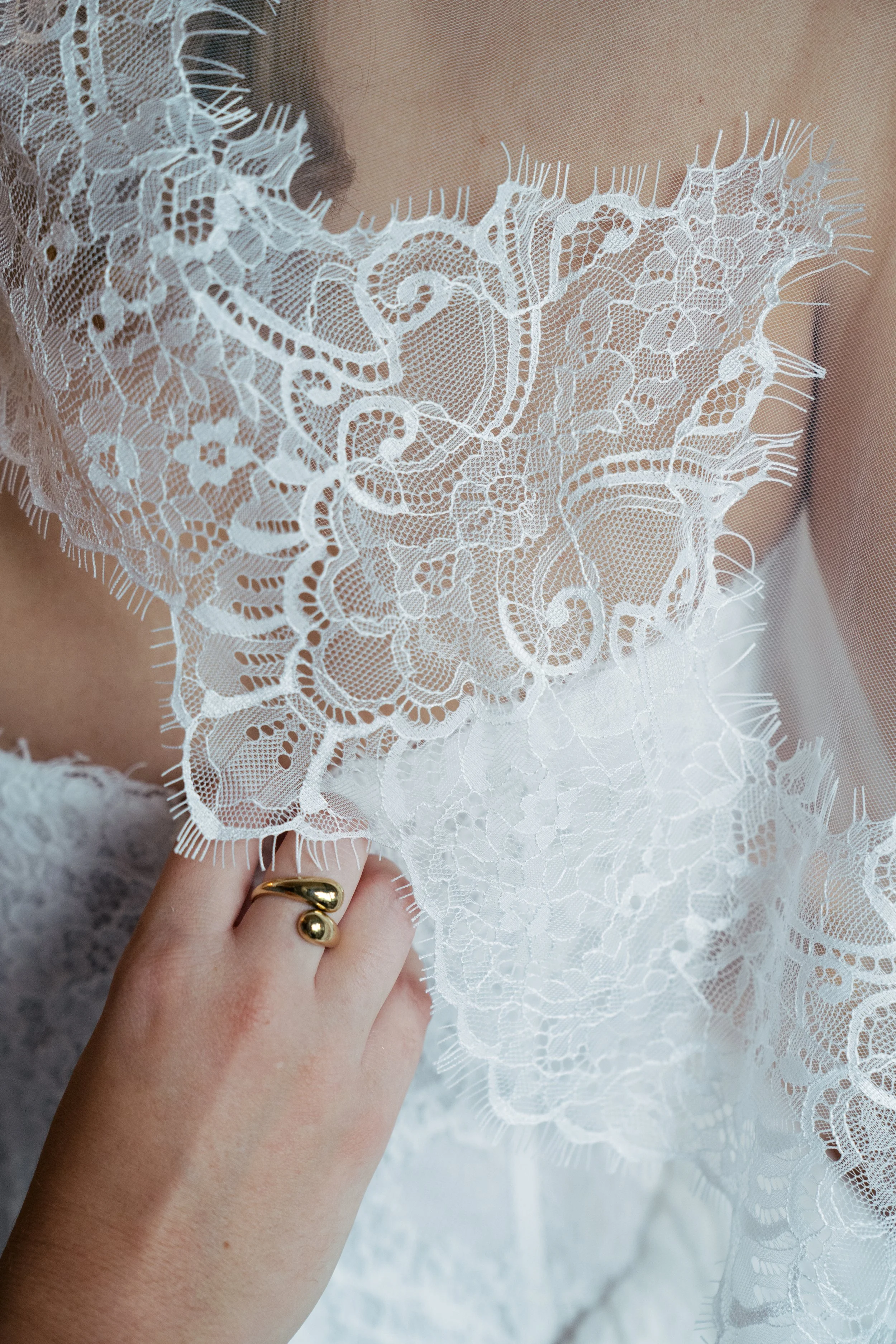 Nahaufnahme eines weißen Spitzenkleides mit floralen Mustern, gehalten von einer Hand mit Goldring.