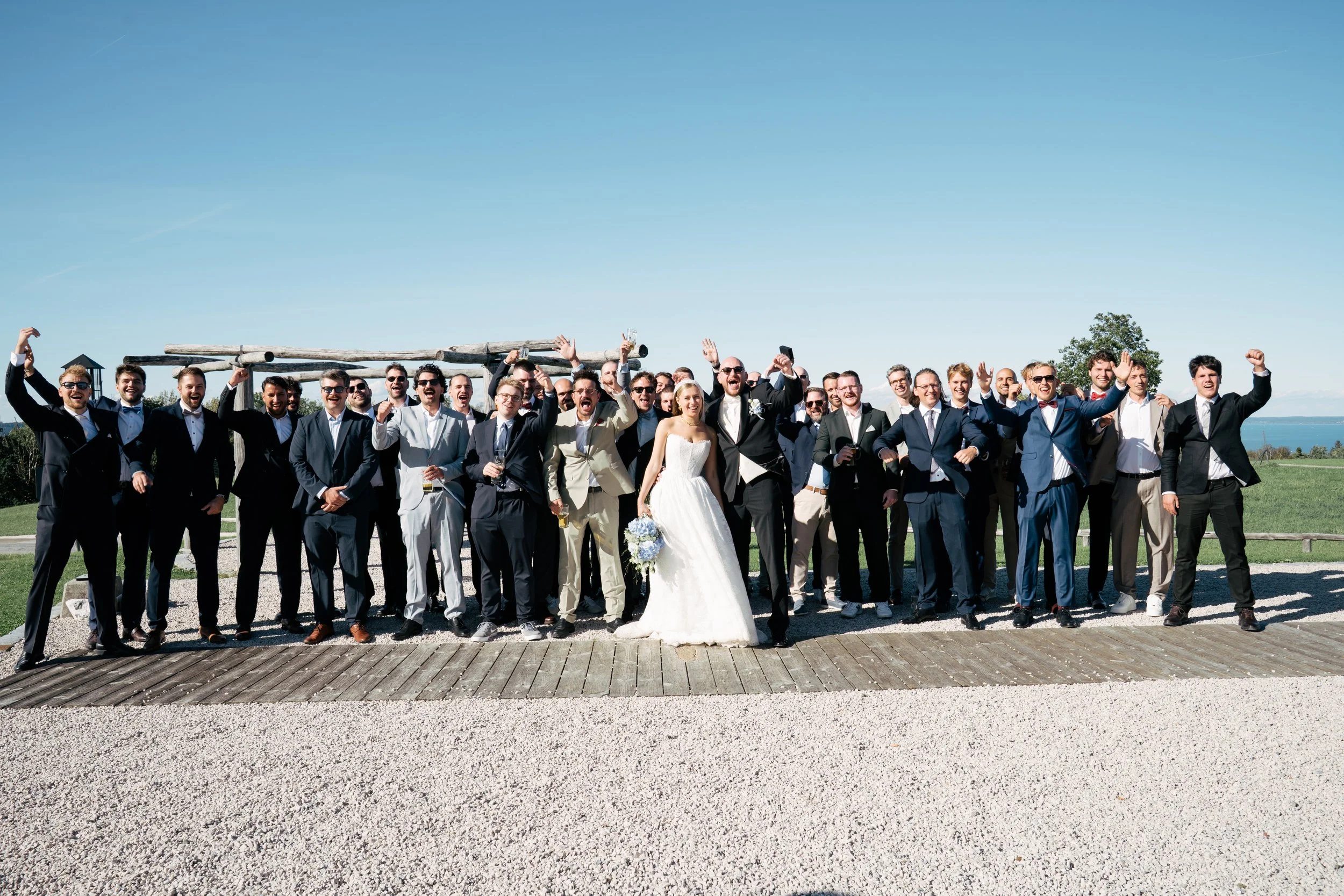 Hochzeitspaar mit Freunden und Familie im Freien, lachend und feiernd, bei sonnigem Wetter mit blauen Himmel im Hintergrund.