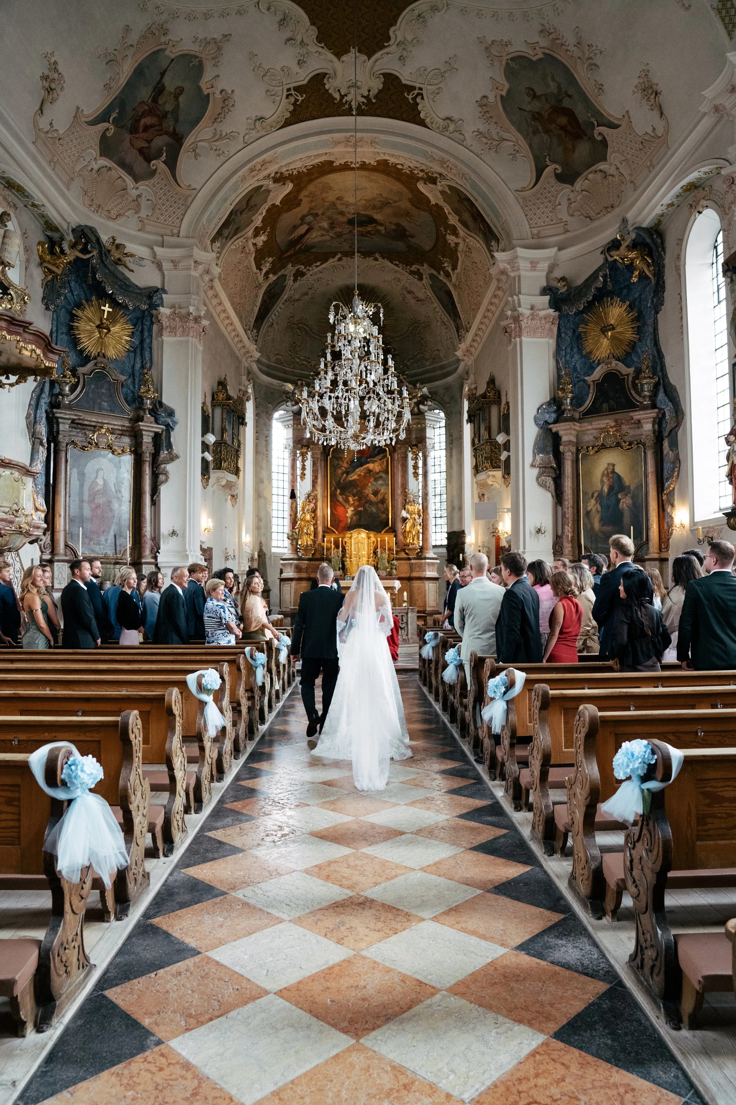 Ein Hochzeitspaar läuft in einer barocken Kirche den Mittelgang entlang, umgeben von Hochzeitsgästen. Die Kirche ist reich verziert mit goldenen Akzenten, Kronleuchtern, Gemälden und kunstvollen Schnitzereien.