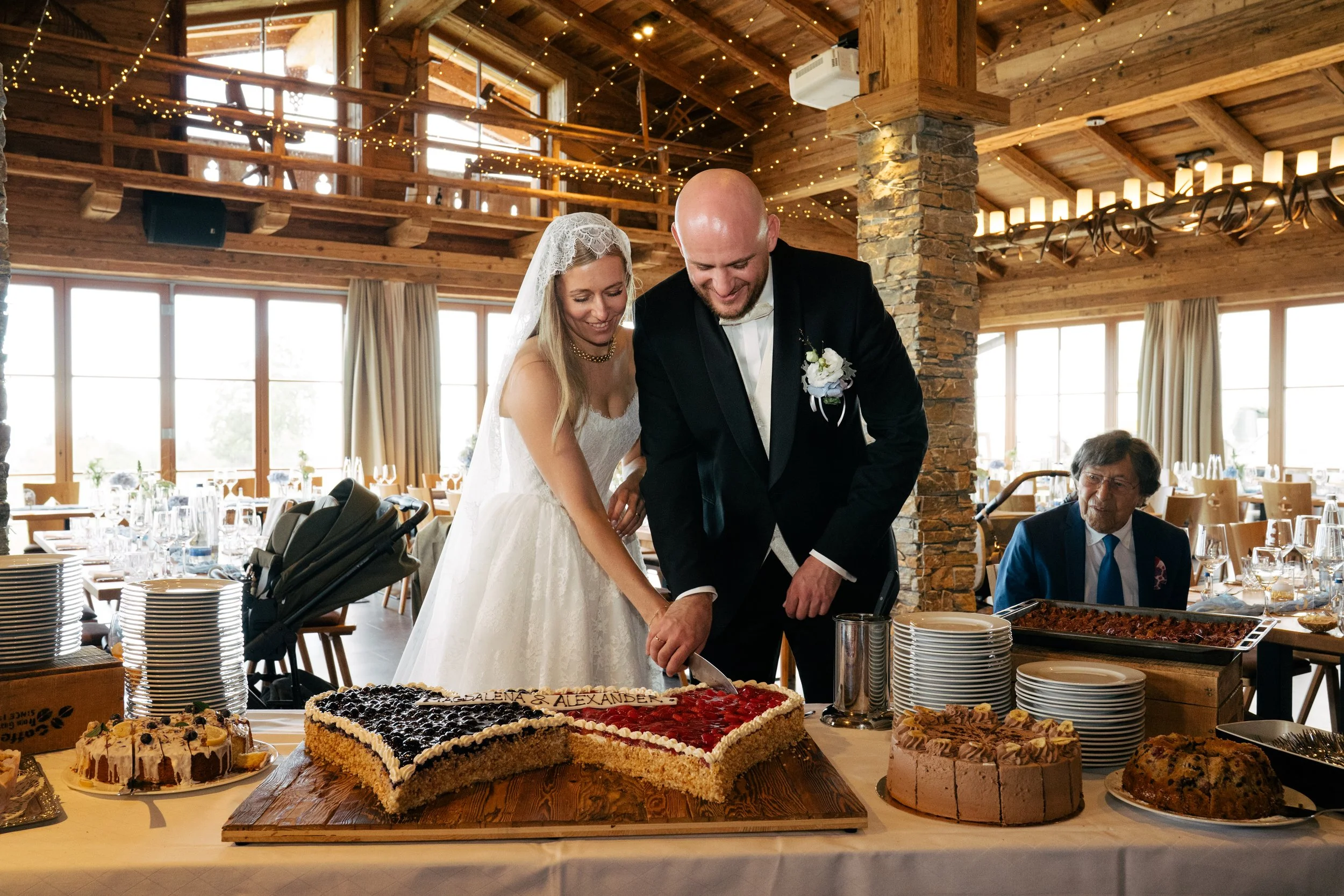 Ein Brautpaar schneidet eine Hochzeitstorte an einem festlichen Empfang in einem rustikalen Raum mit Holzdecke und Dekor aus Lichterketten. Im Vordergrund sind verschiedene Kuchen auf einem Buffet sichtbar.