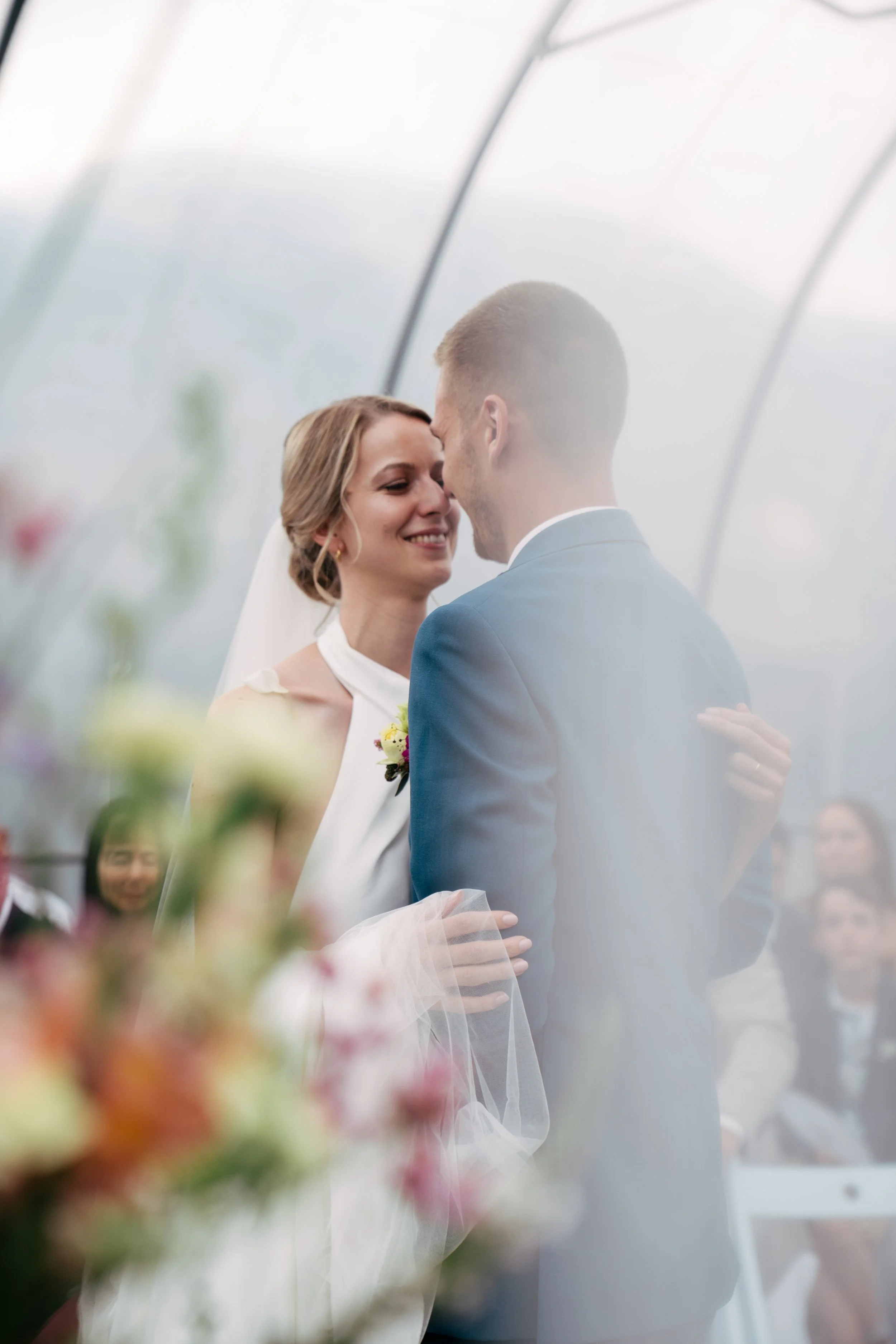 Ein Hochzeitsfoto eines Paares, das sich im Inneren eines Glashauses küsst. Im Vordergrund sind unscharfe Blumen sichtbar.