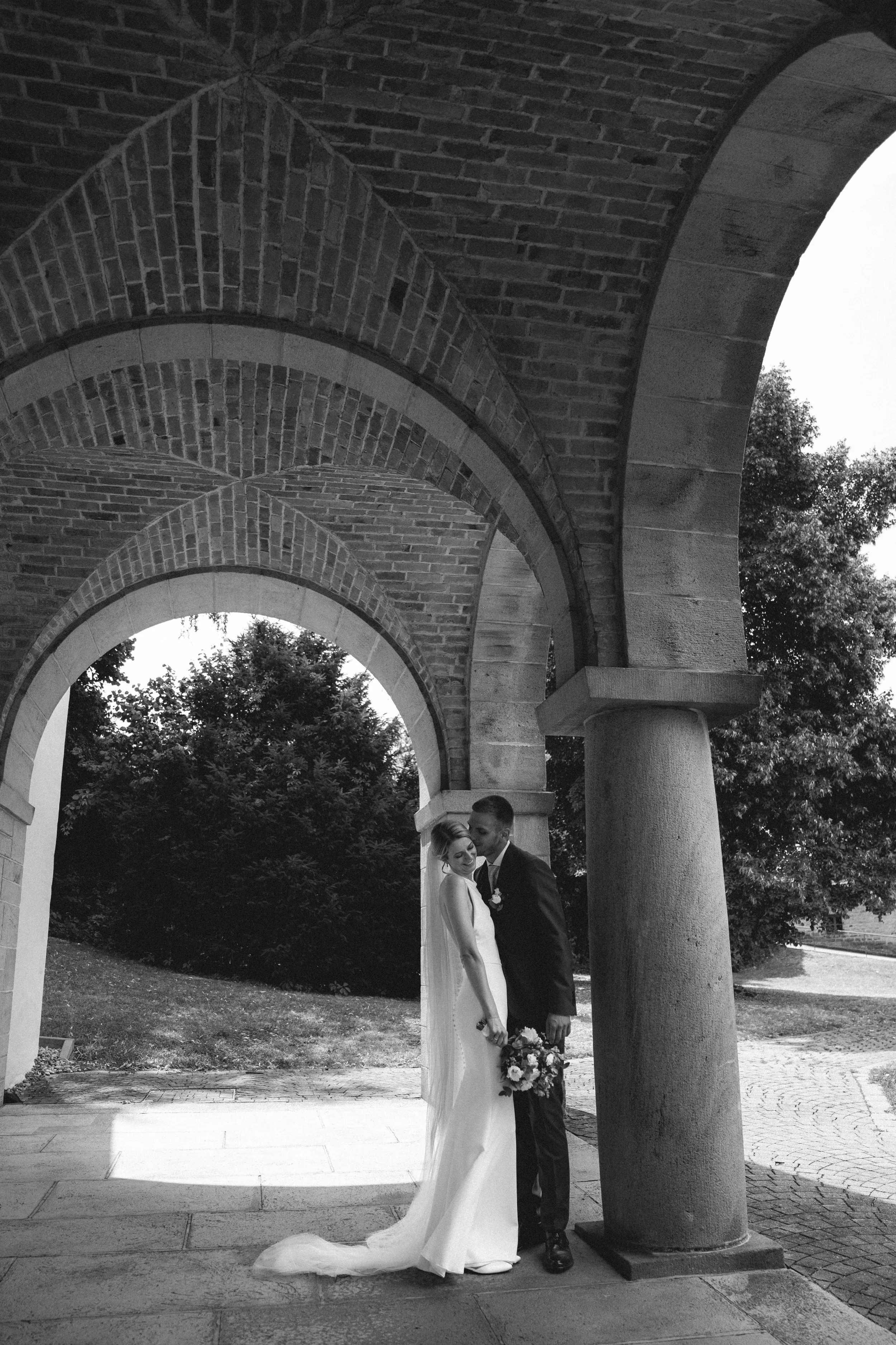 Braut und Bräutigam unter einer Steinbogenbrücke beim Hochzeitsshooting, umgeben von Bäumen, im schwarzen und weißen Foto.