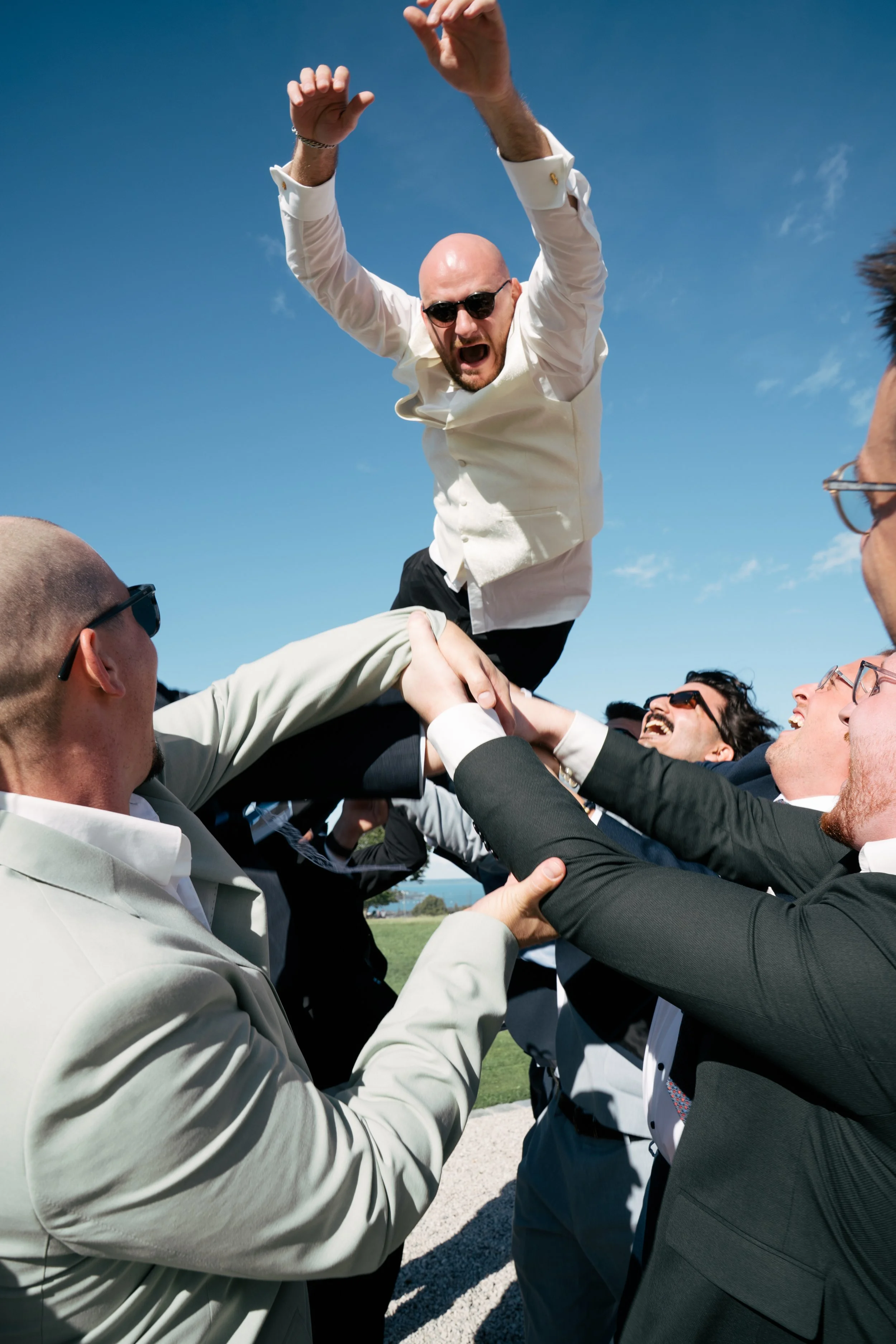 Festgruppe von Menschen, die eine Person auf Schultern tragen, bei einer Feier im Freien, blauer Himmel, einige tragen Sonnenbrillen, fröhliche Stimmung.