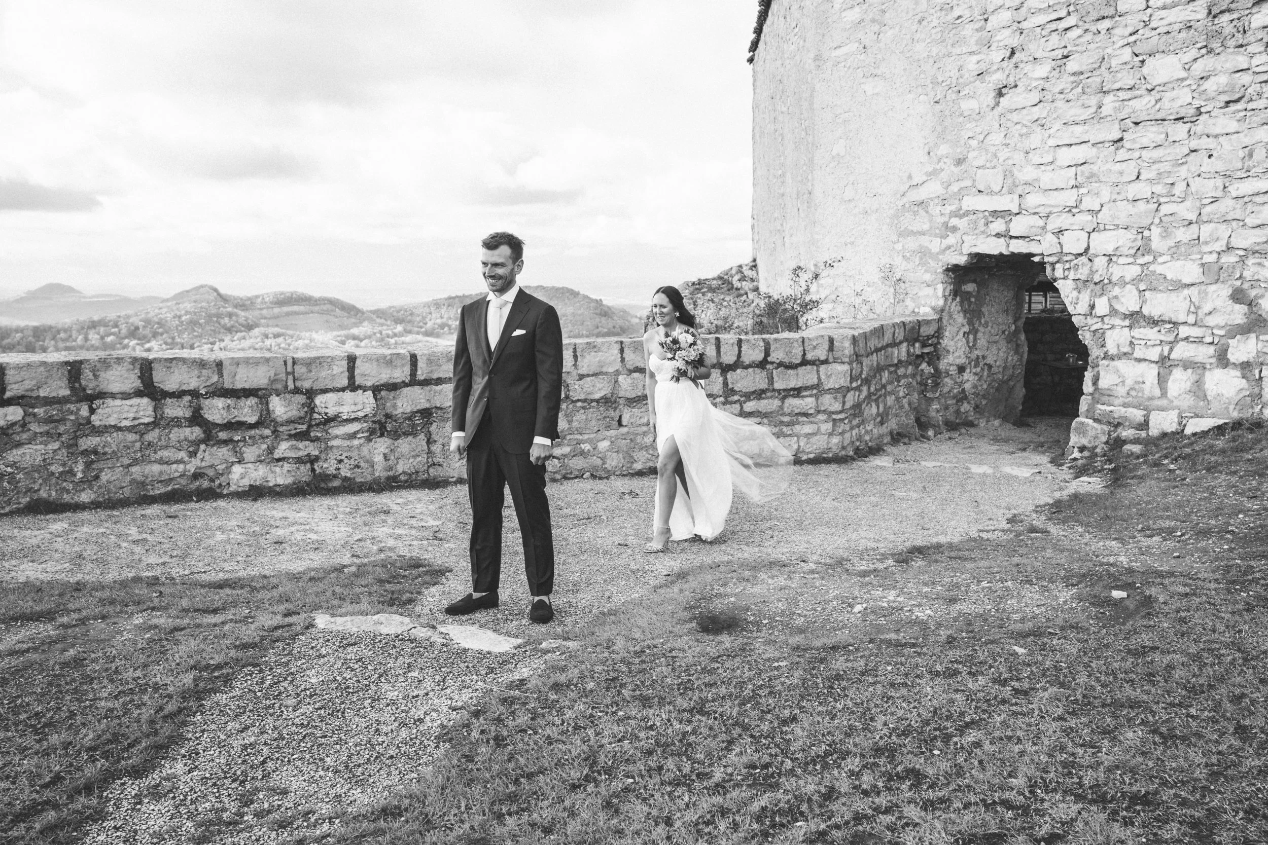 Schwarzweißfoto von einem Paar draußen, der Mann im Anzug steht vorne, die Frau im Brautkleid geht von hinten lächelnd auf ihn zu. Sie hält einen Blumenstrauß. Im Hintergrund sind eine alte Steinmauer und sanfte Hügel zu sehen.