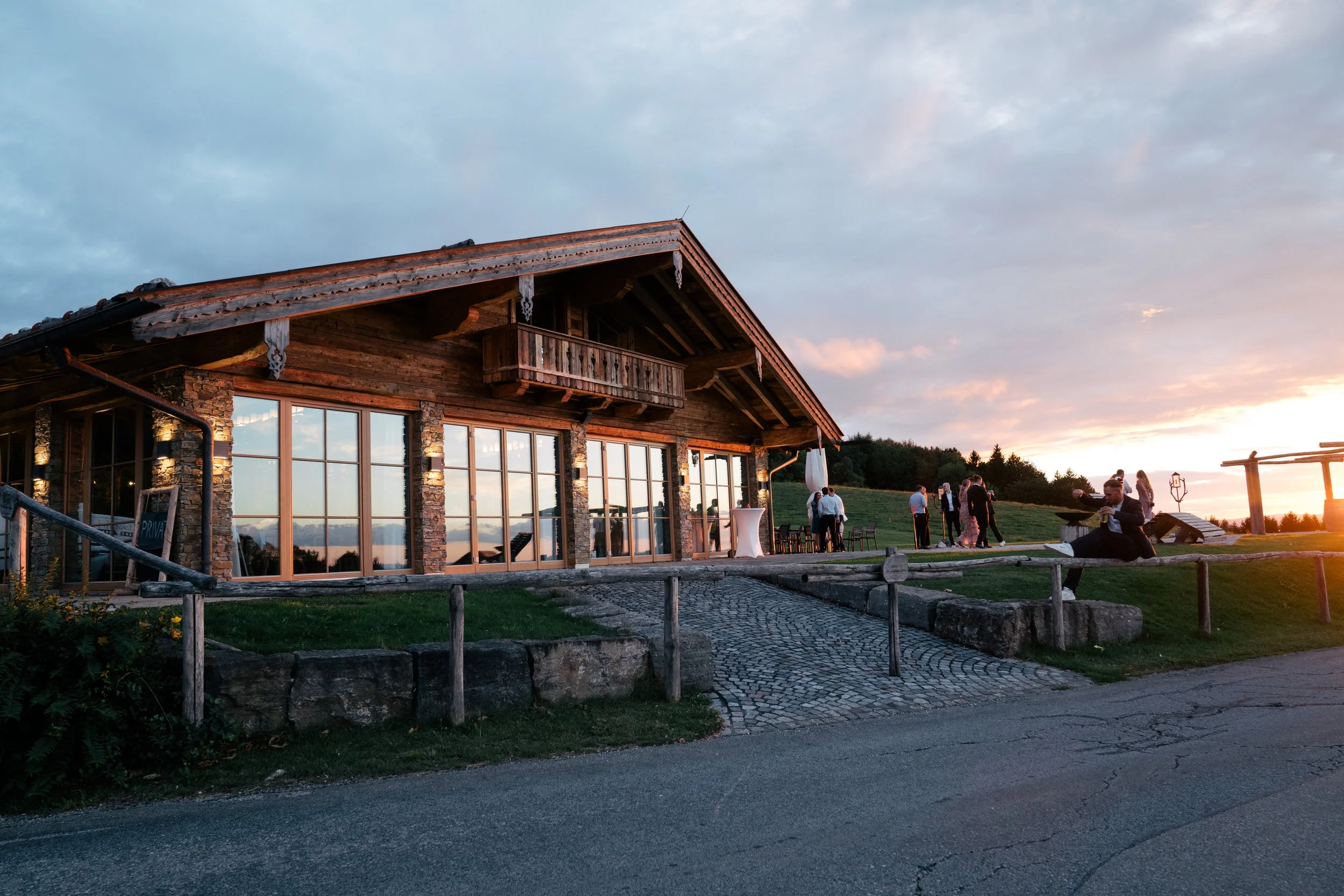 Ein rustikales Holzhaus mit großen Glasfenstern und einem Balkon unter einem schieferbedeckten Dach bei Sonnenuntergang, mit mehreren Personen im Freien, die sich unterhalten und bei einer Veranstaltung im Grünen am Abend