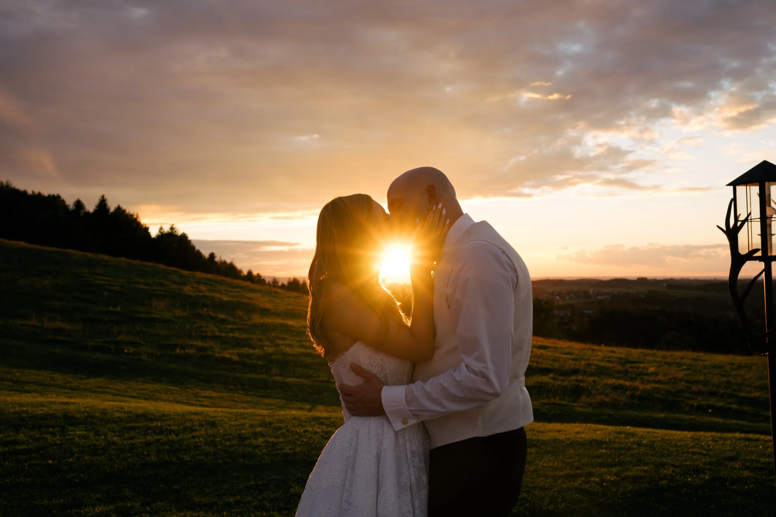 Ein Brautpaar umarmt sich bei Sonnenuntergang im Freien, die Frau trägt ein weißes Kleid, der Mann ein weißes Hemd, im Hintergrund sind Bäume und eine Landschaft mit einer alten Wetterfahne.