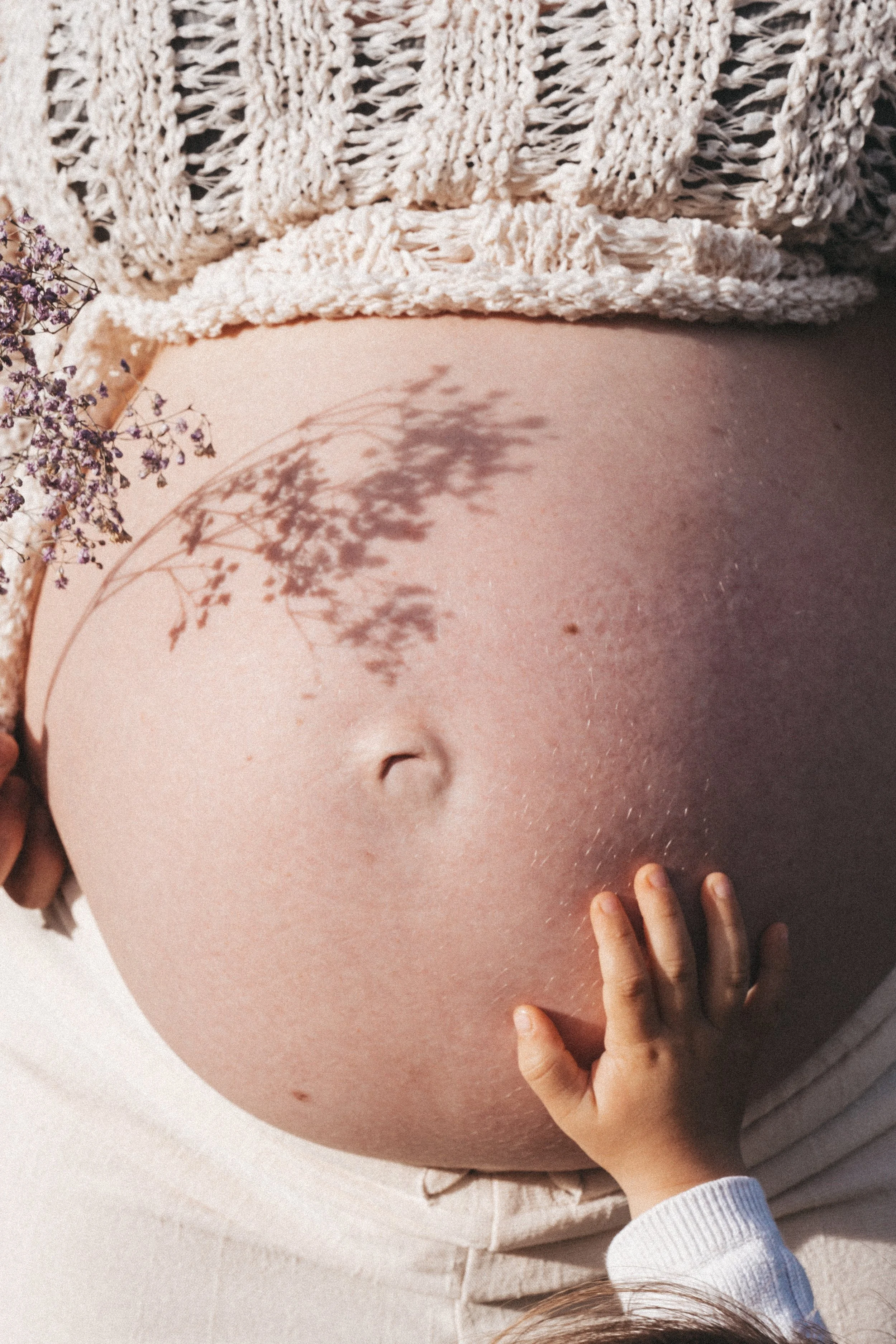 Nahaufnahme eines schwangeren Bauches mit einem kleinen Kinderarm, der ihn berührt, neben einem Blumenstrauß und einem handgeschriebenen Schatten. Die Szene wird durch warmes Licht beleuchtet.