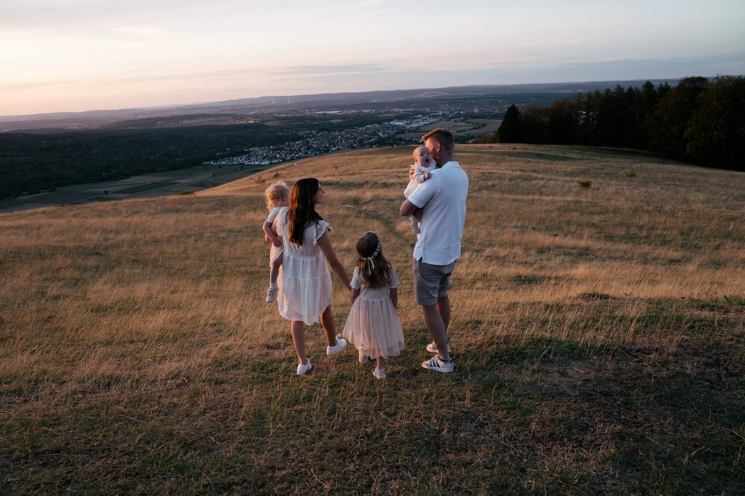 Familie mit drei Kindern und einem Mann steht auf einem Hügel bei Sonnenuntergang und schaut auf die Landschaft, die sich vor ihnen erstreckt, mit Wäldern und einer Stadt im Hintergrund.