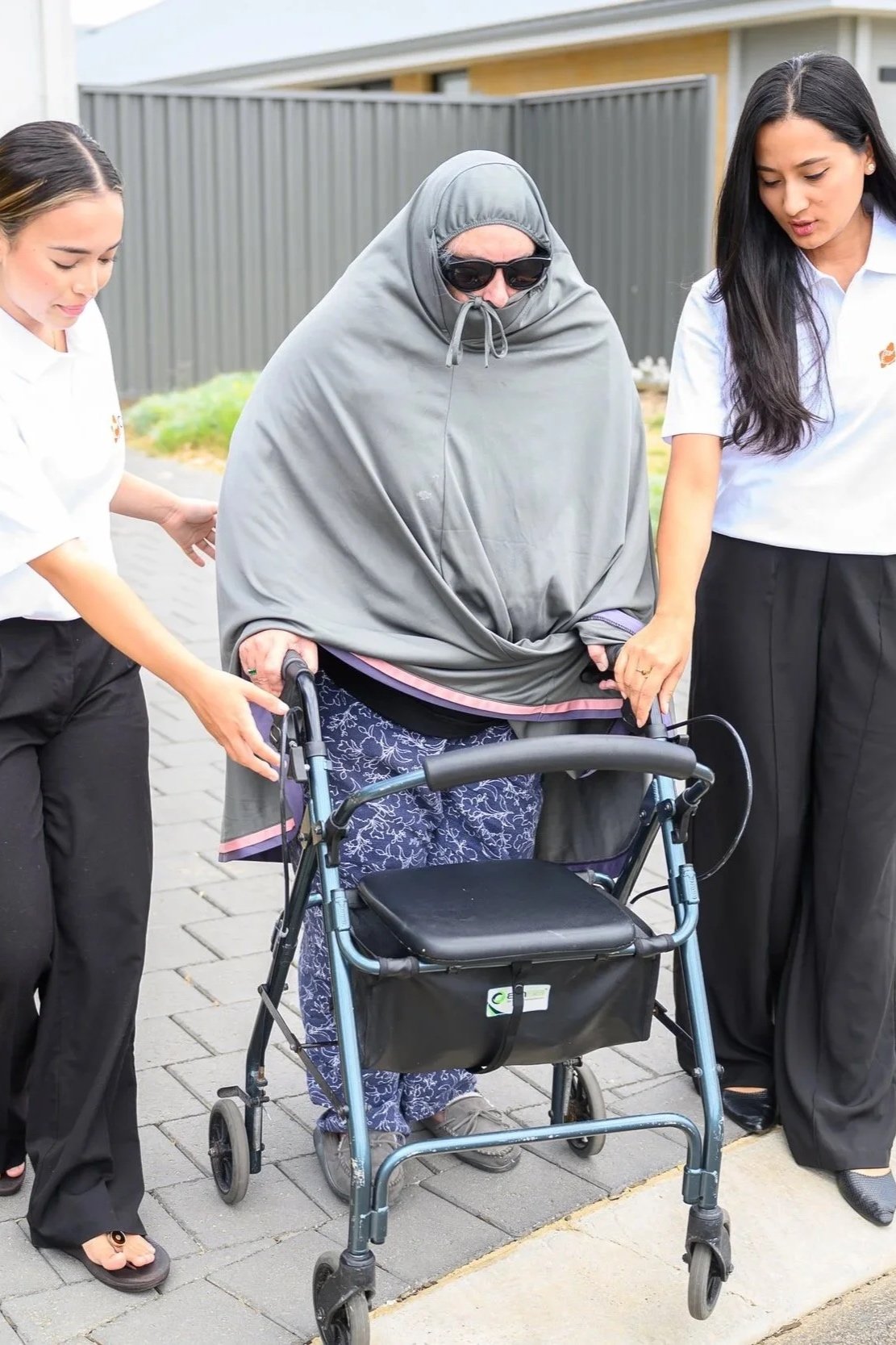 An elderly woman wearing sunglasses and a gray hijab, using a walker, being assisted by two young women in white shirts, outside near a fence.