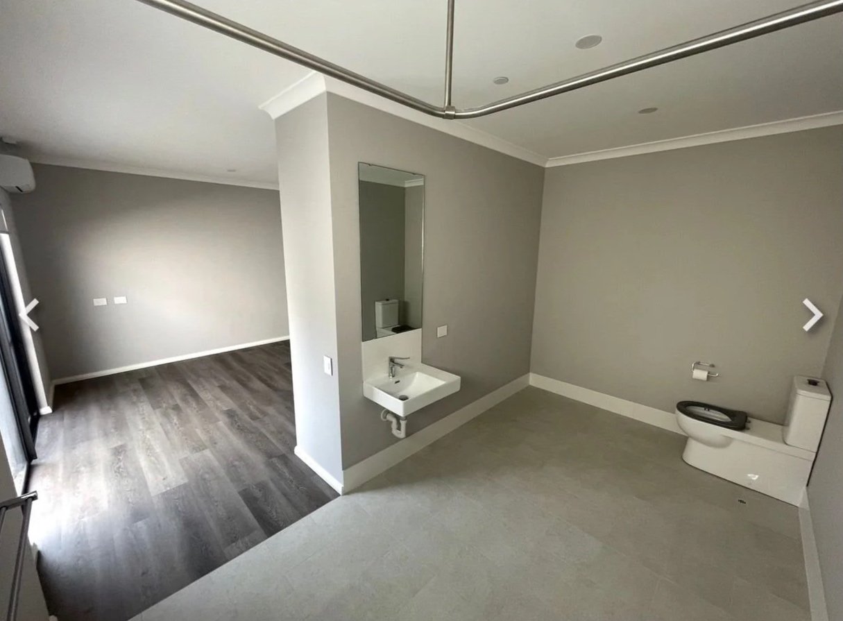 Unfinished bathroom with a small sink, mirror, and toilet, adjacent to a living area with dark wood flooring, cloudy windows, and gray walls.