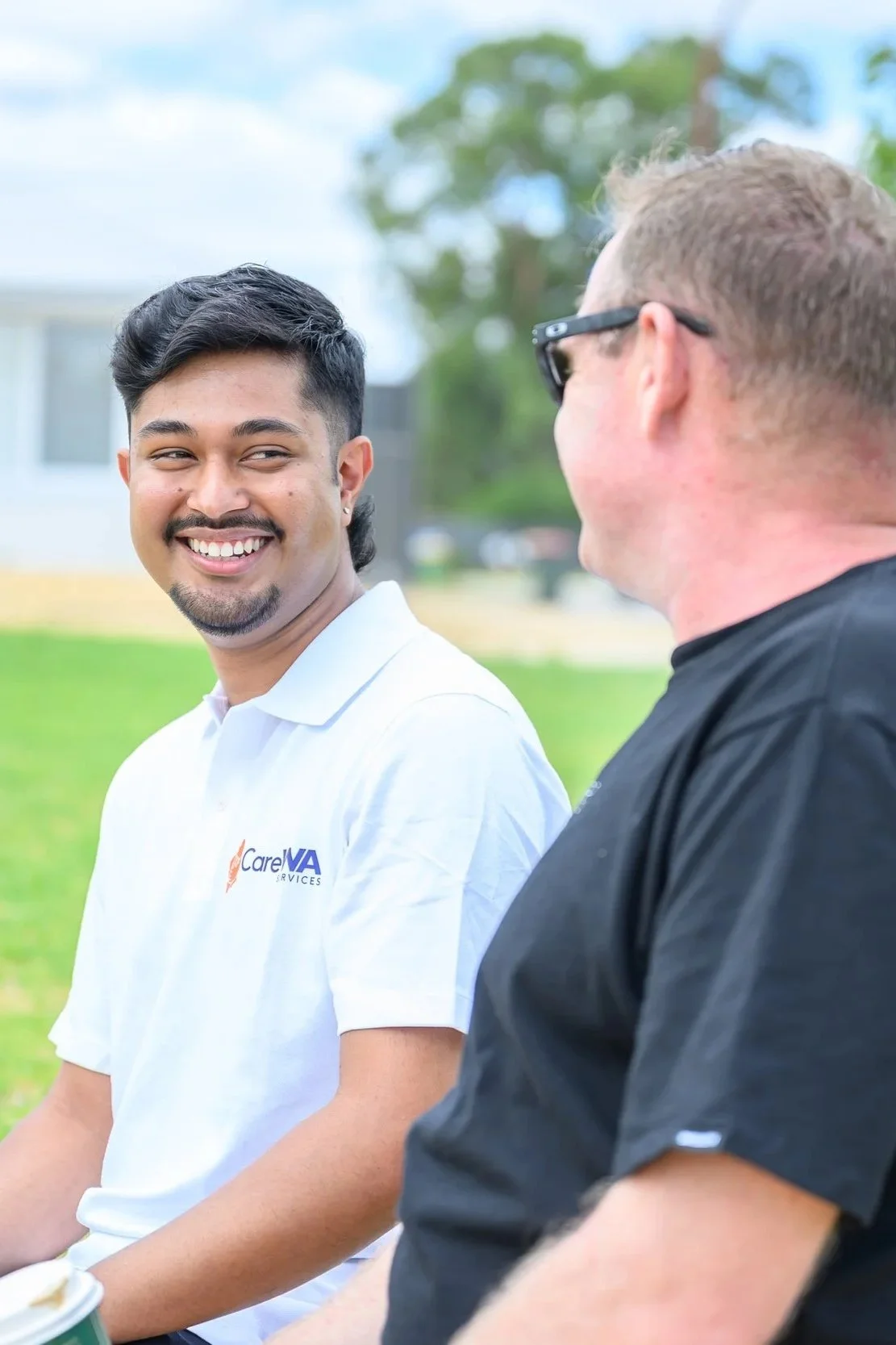 Two men sitting outside on a sunny day, having a conversation. The man on the left is smiling and wearing a white polo shirt with a CareVA logo. The man on the right is wearing sunglasses and a black shirt, and is looking at the other man.