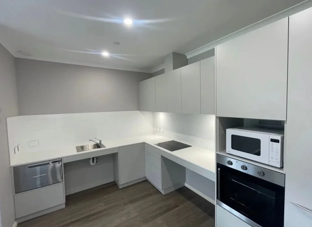 Modern kitchen with white cabinets, stainless steel sink, electric stovetop, microwave, and oven, with gray walls and wood flooring.