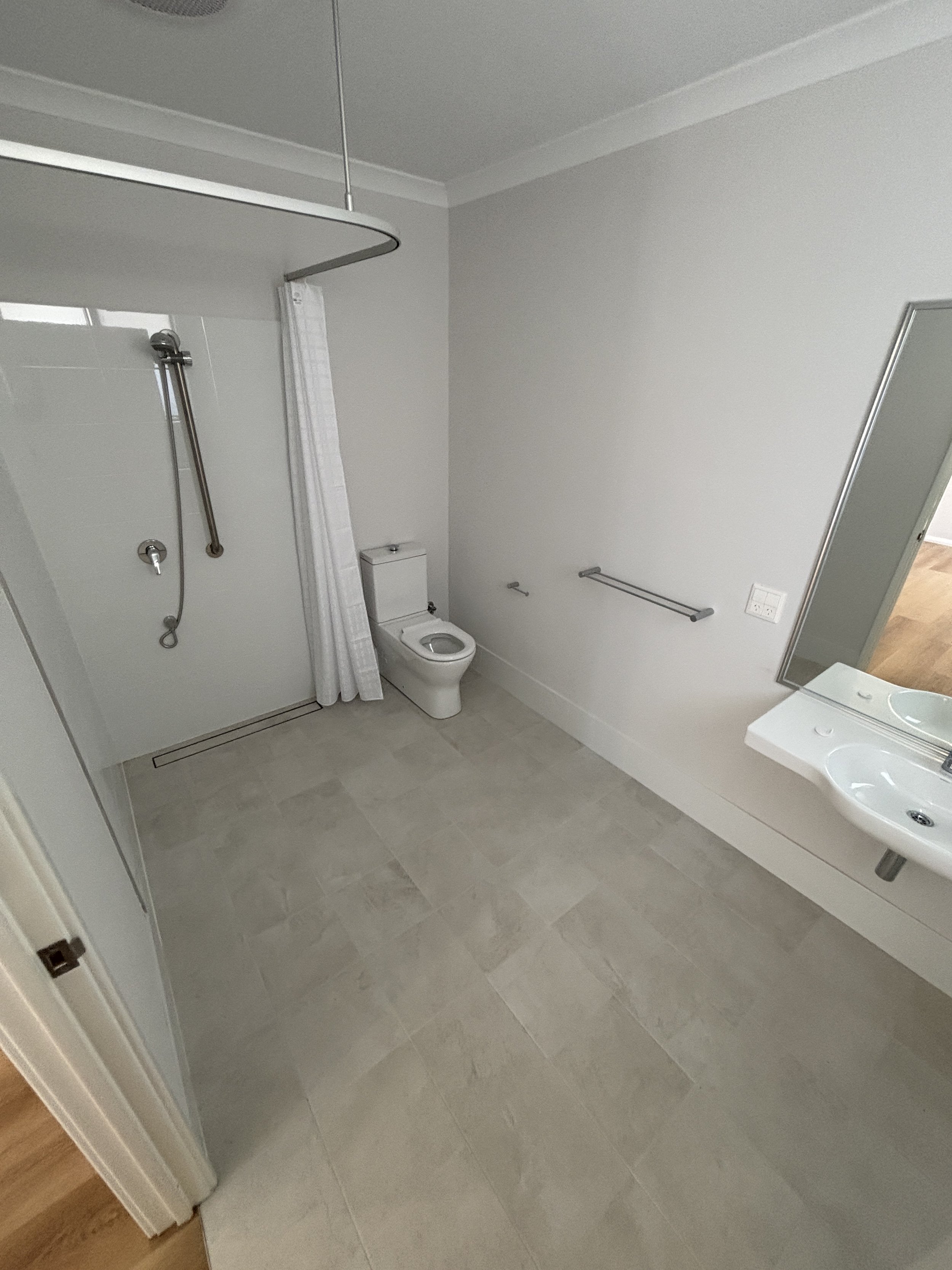 An empty bathroom with a walk-in shower, a toilet, a sink with a large mirror, and towel bars on the wall. The bathroom has light-colored tiled flooring and white walls.