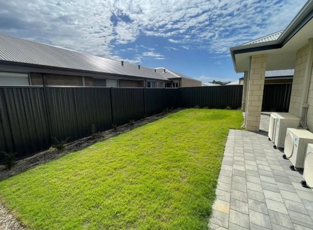 Backyard with a green lawn, paved patio area with air conditioning units, black fence, and row of houses with metal roofs under a partly cloudy sky.