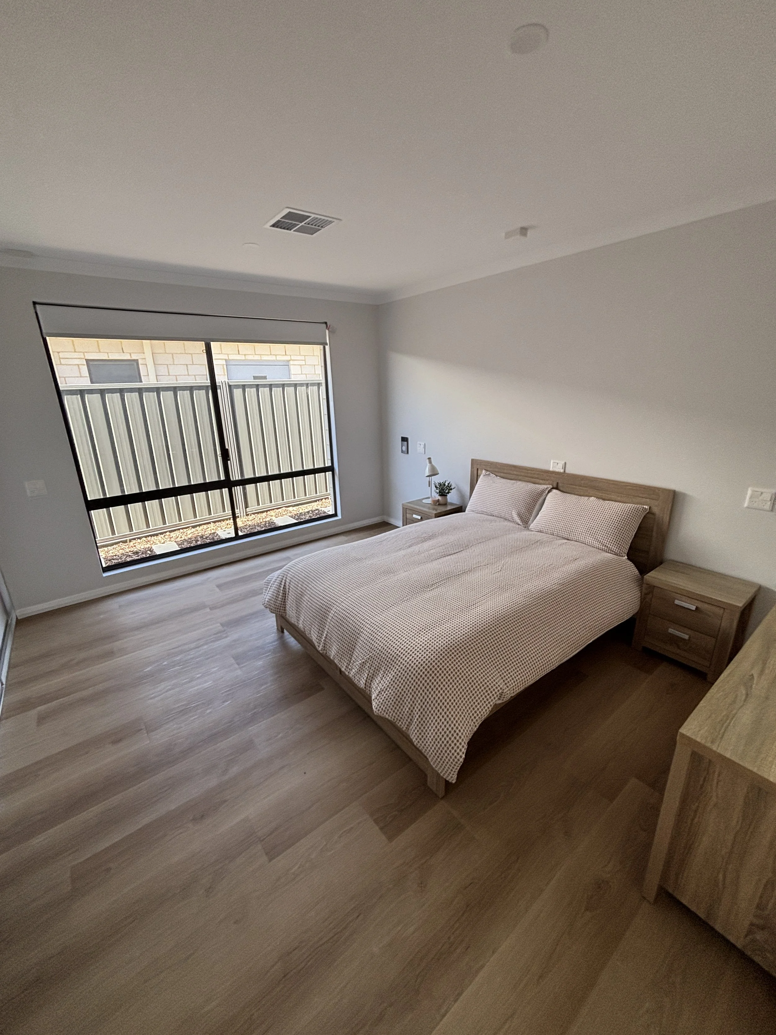 A bedroom with a bed, two nightstands, a window with blinds, a small lamp, and a potted plant, featuring light-colored wooden flooring and white walls.