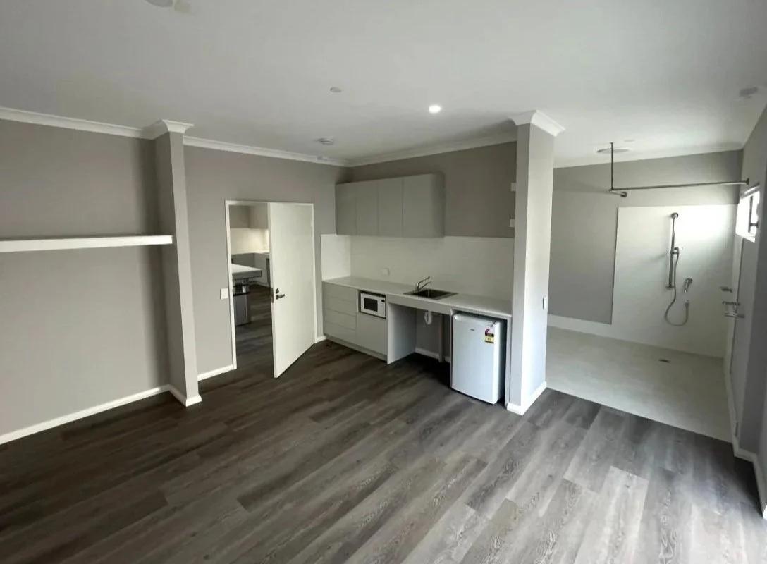 Empty apartment with an open kitchen, including a small refrigerator, microwave, and sink, adjacent to a laundry area with plumbing and a shower, all with gray walls and wood laminate flooring.