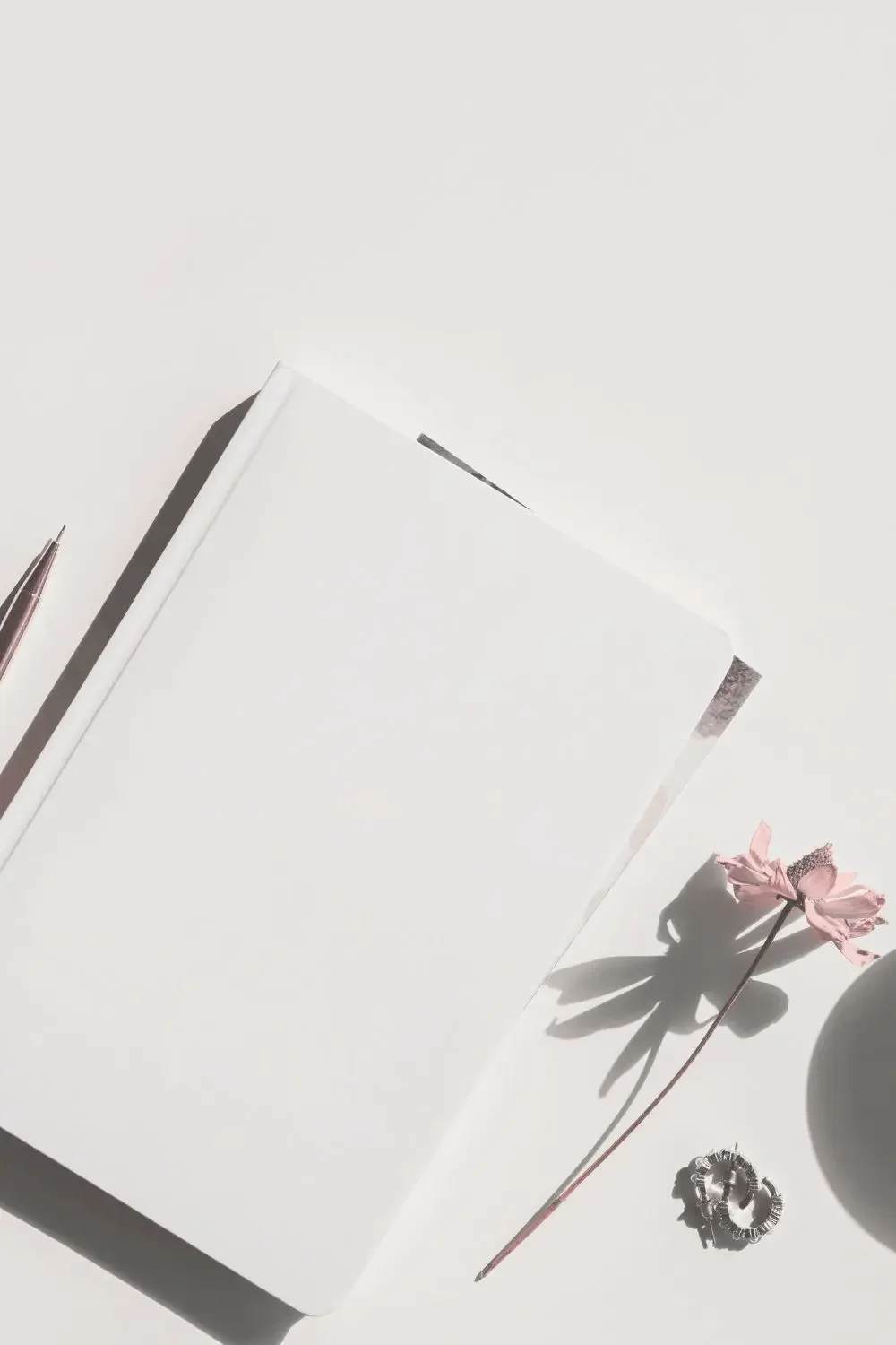 Ein weißes notizbuch, ein rosa Blumenstängel, ein Kugelschreiber und eine silberne Haarklammer liegen auf einem weißen Tisch.