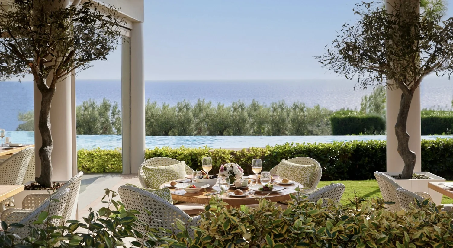 A table set for lunch on a shaded terrace overlooking an infinity pool and the sea