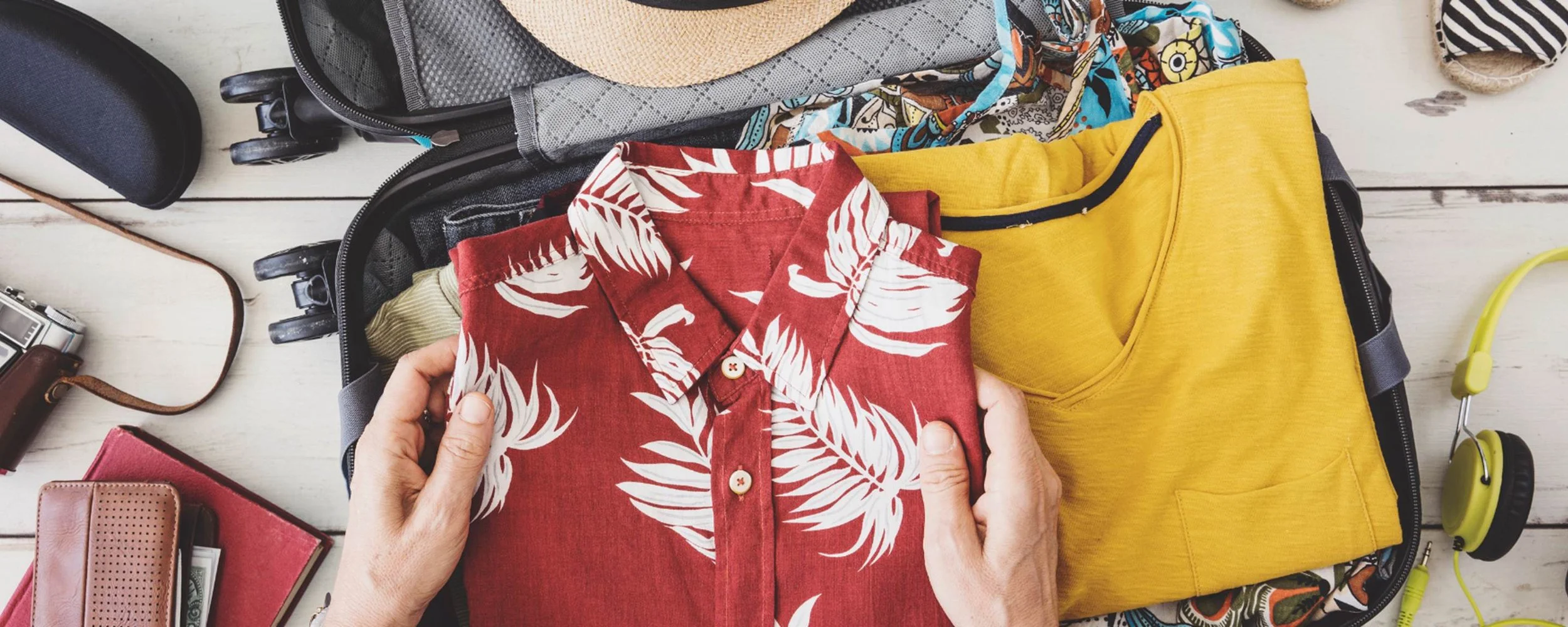 Hands holding a folded red tropical print shirt over an open suitcase with a camera and headphones