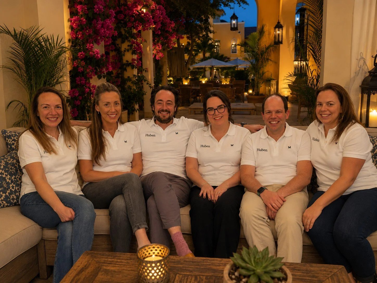 The six members of the Huben Travel team in white Huben polo shirts, sitting together on an outdoor sofa at dusk
