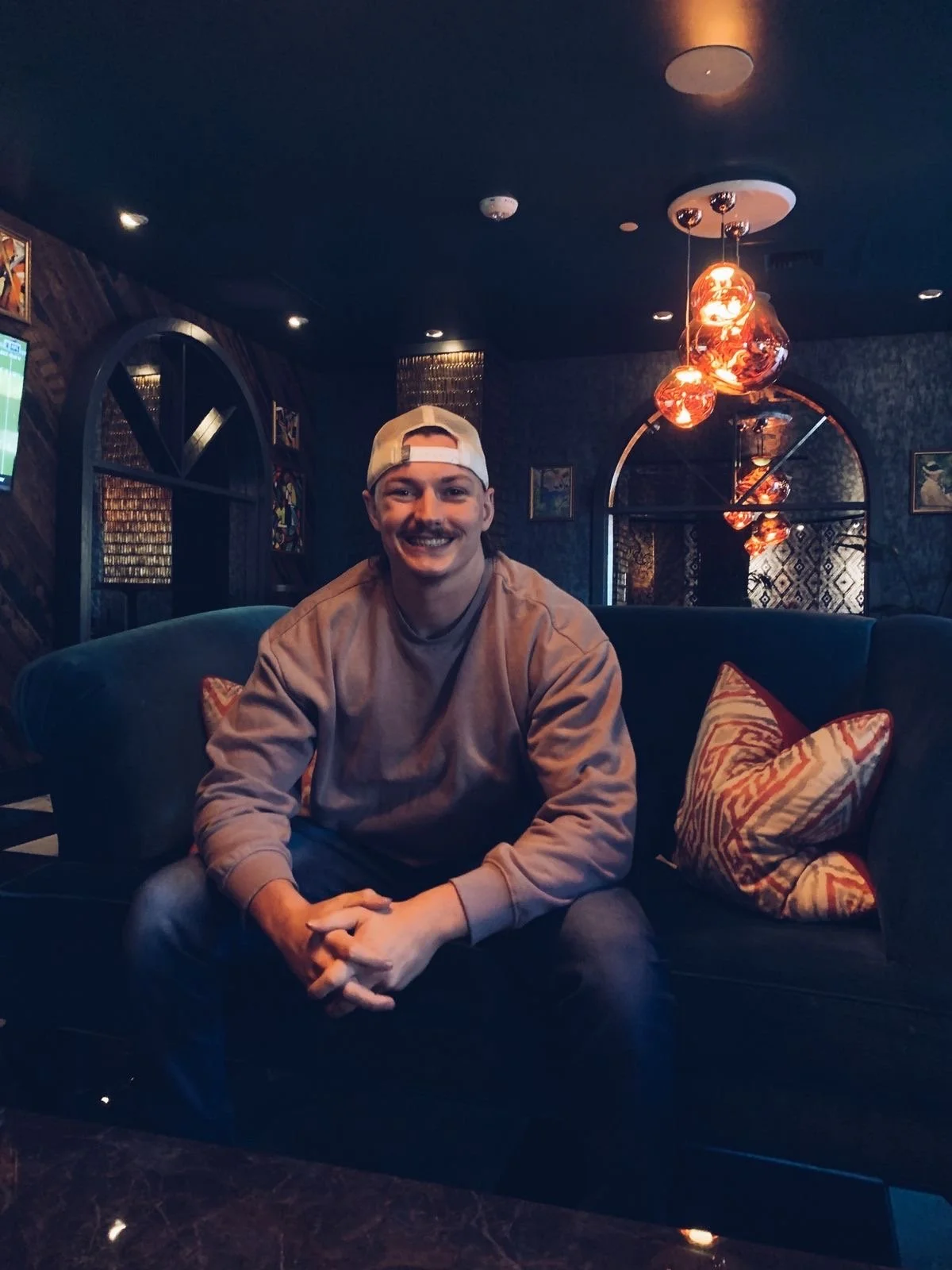 A young man sitting on a dark blue sofa in a dimly lit room, smiling at the camera, wearing a beige baseball cap backwards and a tan hoodie, with patterned pillows behind him and decorative hanging lights above.