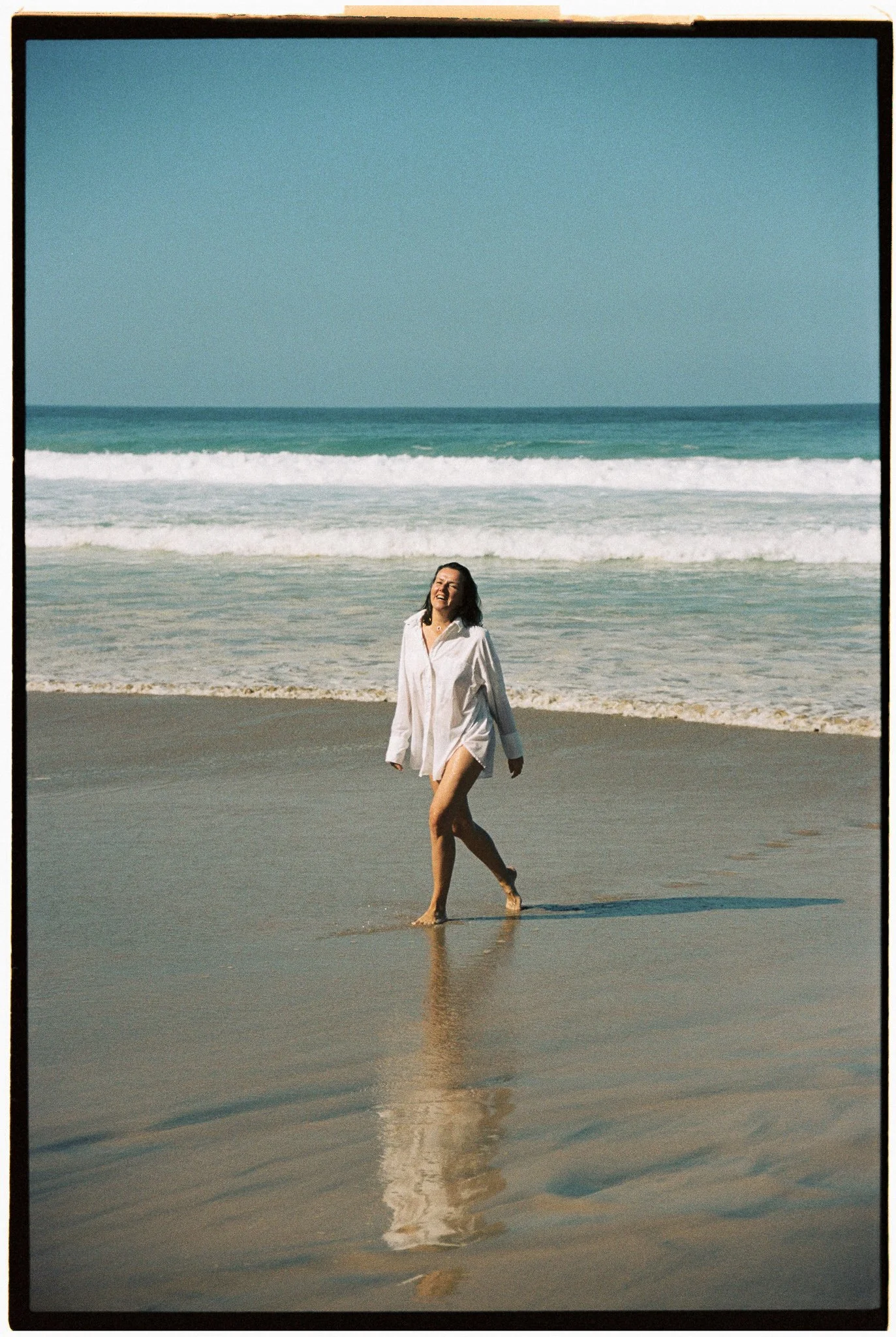 Woman walking on the beach in a white shirt with waves in the background.