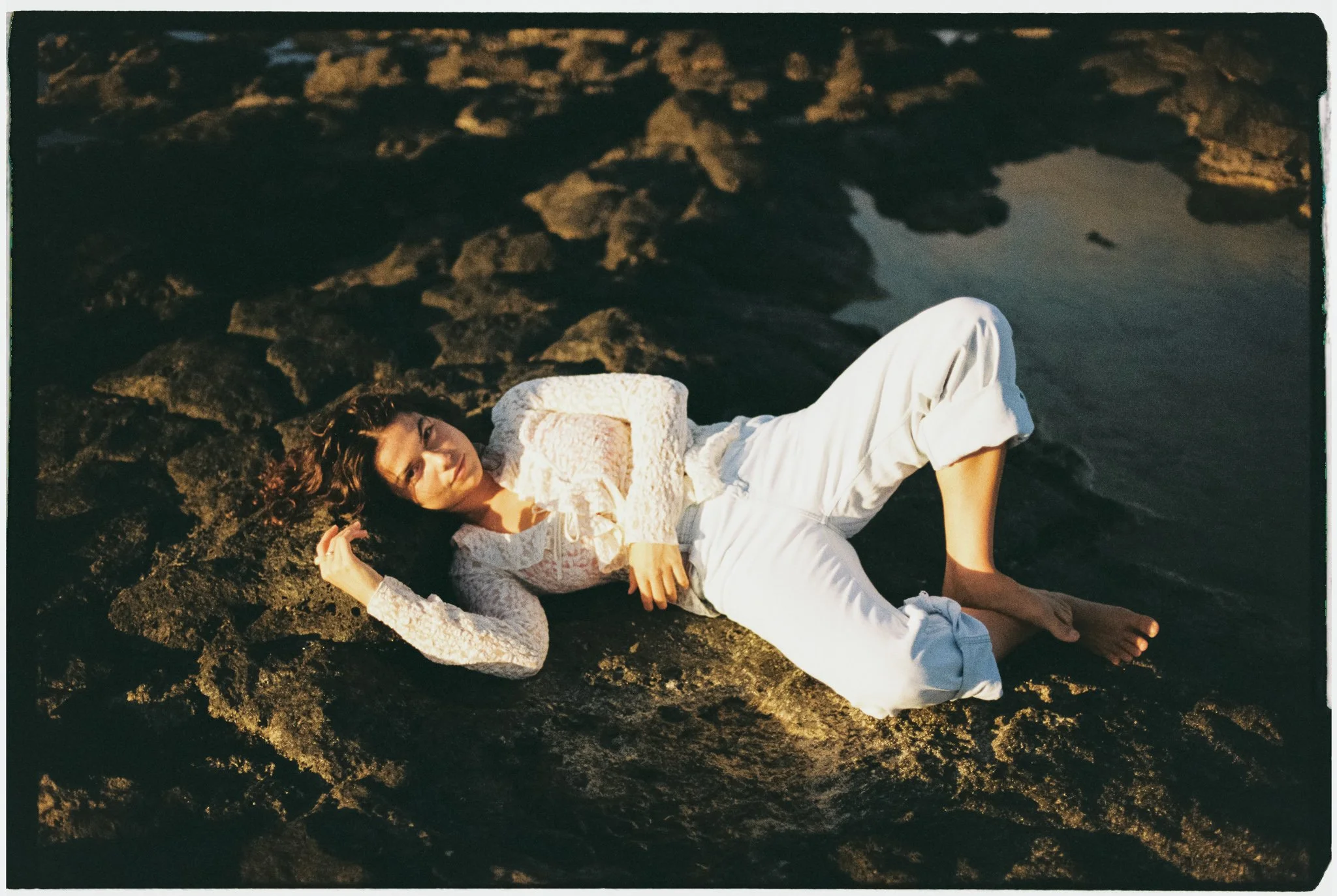 Woman in white lace top and white pants lying on rocky shoreline near water at sunset.