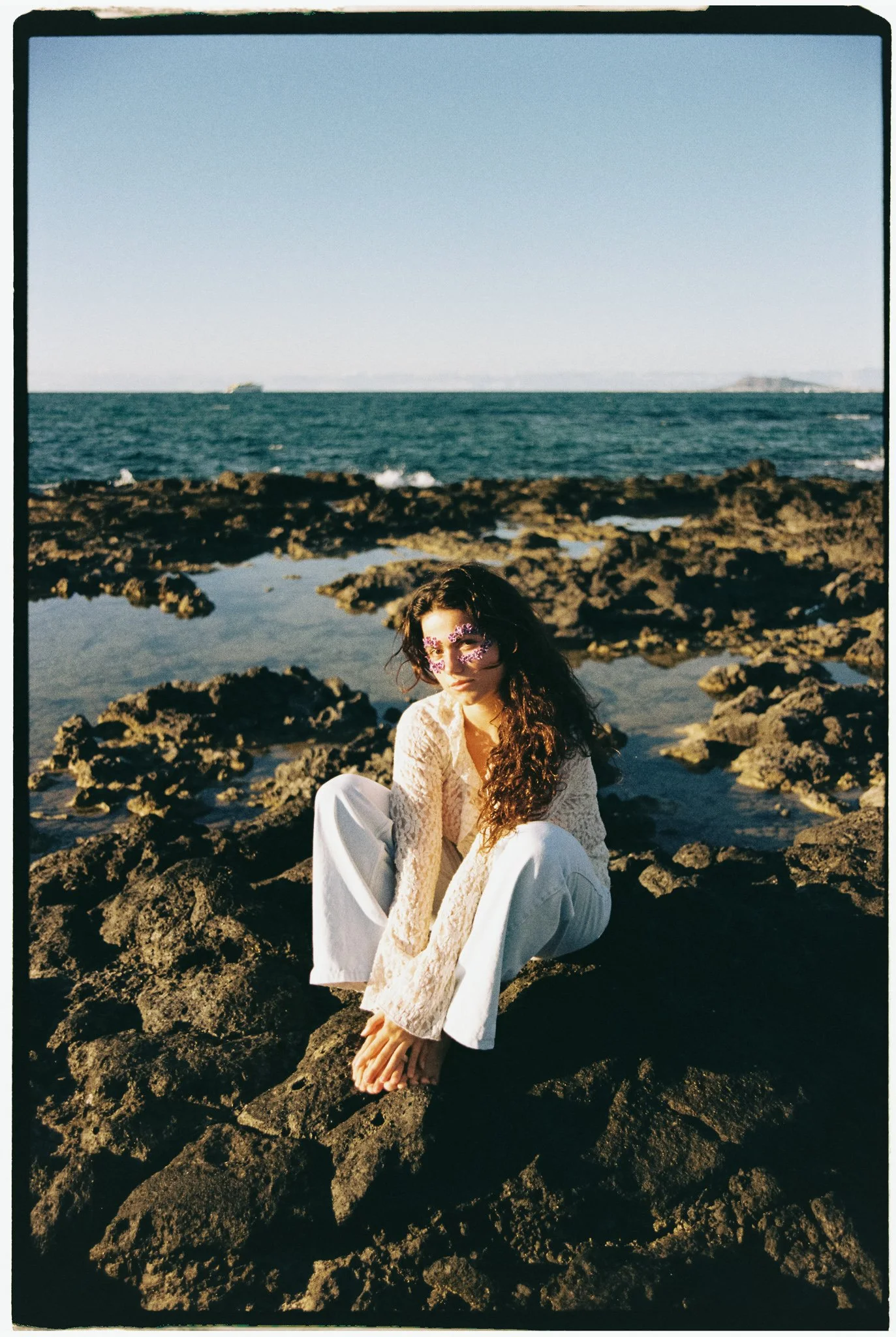 A woman with long curly hair and floral face jewelry crouches on dark volcanic rocks near the ocean, wearing a lace top and wide-leg pants, with the sea and a clear sky in the background.