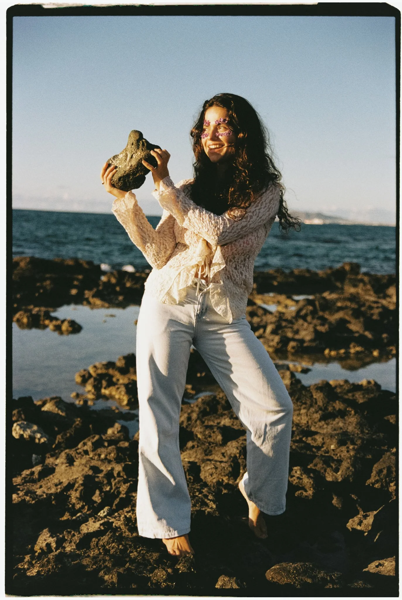 A woman with long curly hair wearing a pink lace long-sleeve top and white pants standing barefoot on rocky beach holding a large piece of volcanic rock in her hands, smiling with the ocean and distant land in the background during sunset.