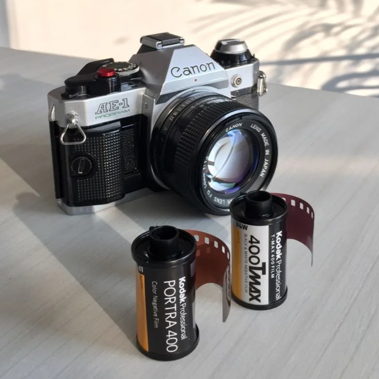 A vintage Canon AE-1 film camera with a lens attached, along with two rolls of Kodak Portra 400 film placed on a light-colored surface with a shadow pattern in the background.