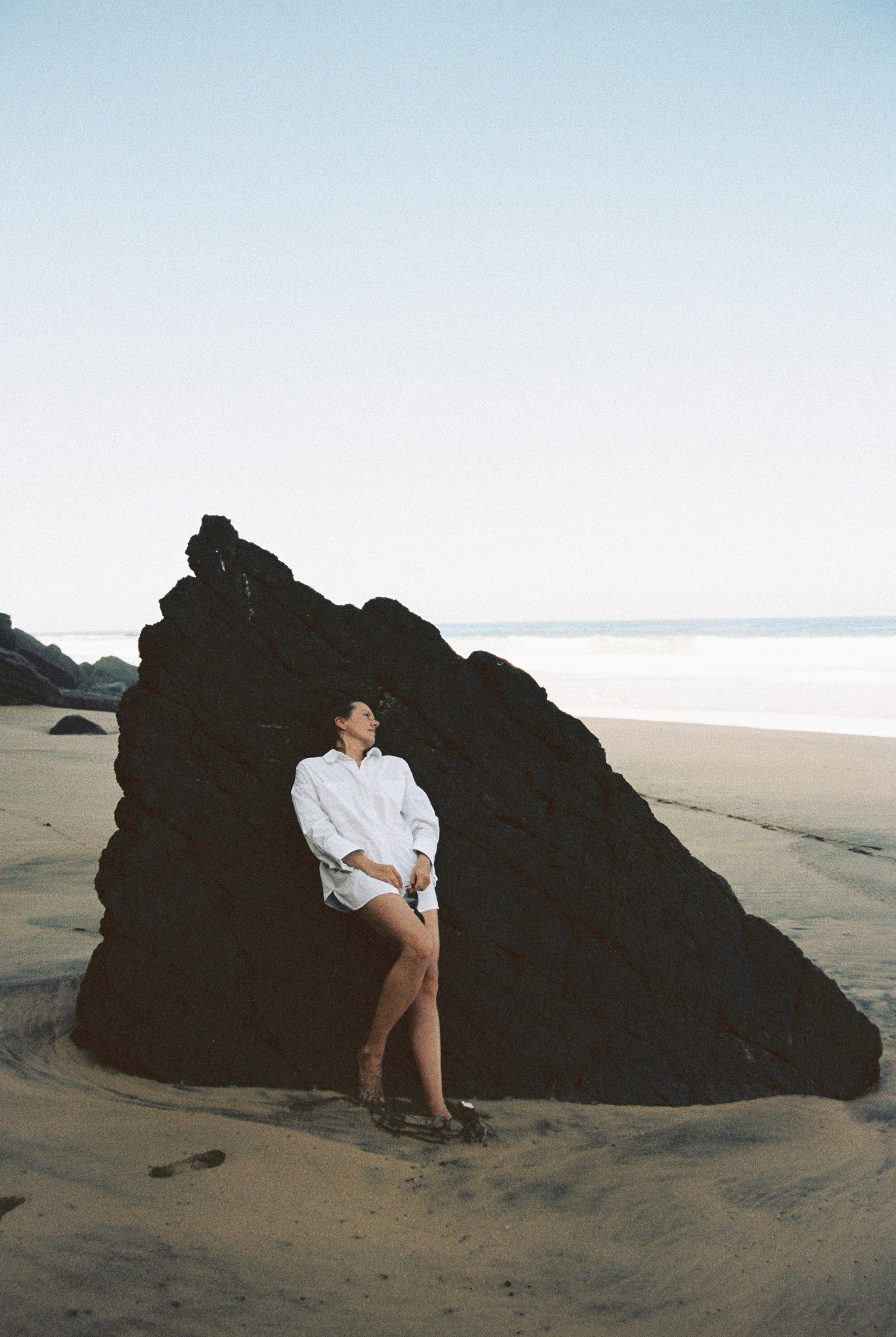 A woman in a white shirt and black sandals leaning against a large black rock on a sandy beach.