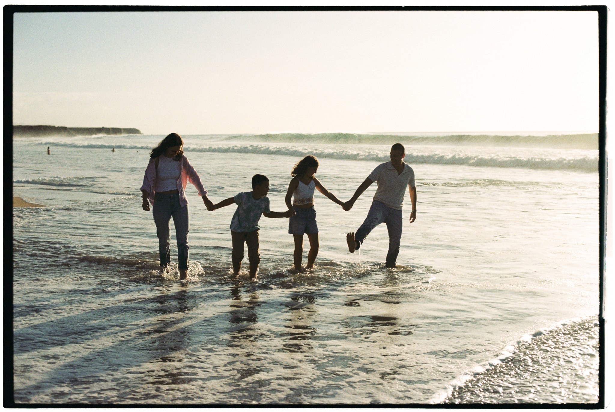 Family of four holding hands and walking in the ocean water at the beach during sunset.