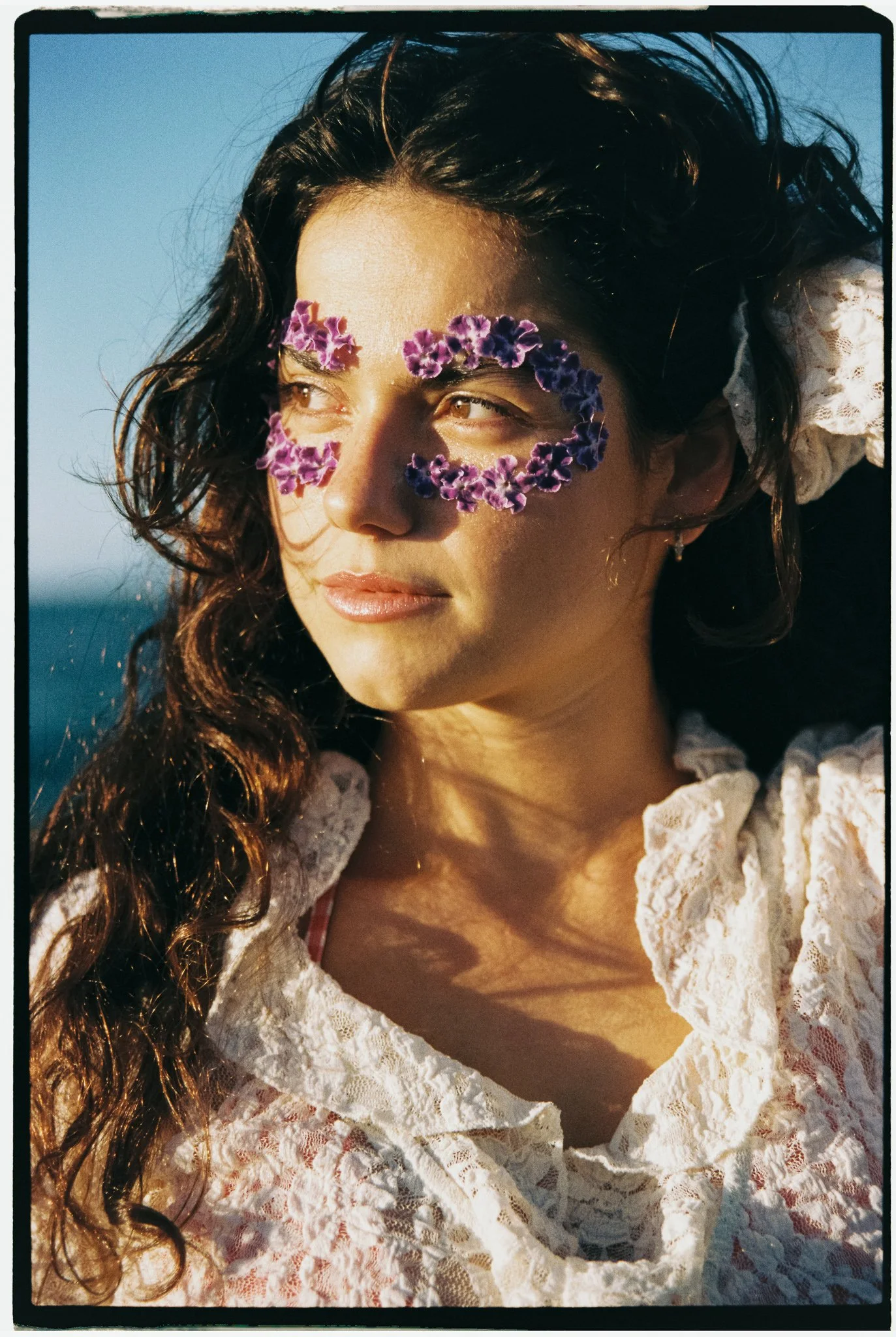 Young woman with curly brown hair wearing glasses made of purple flowers, looking to the side, with ocean in the background, dressed in a white lace top.
