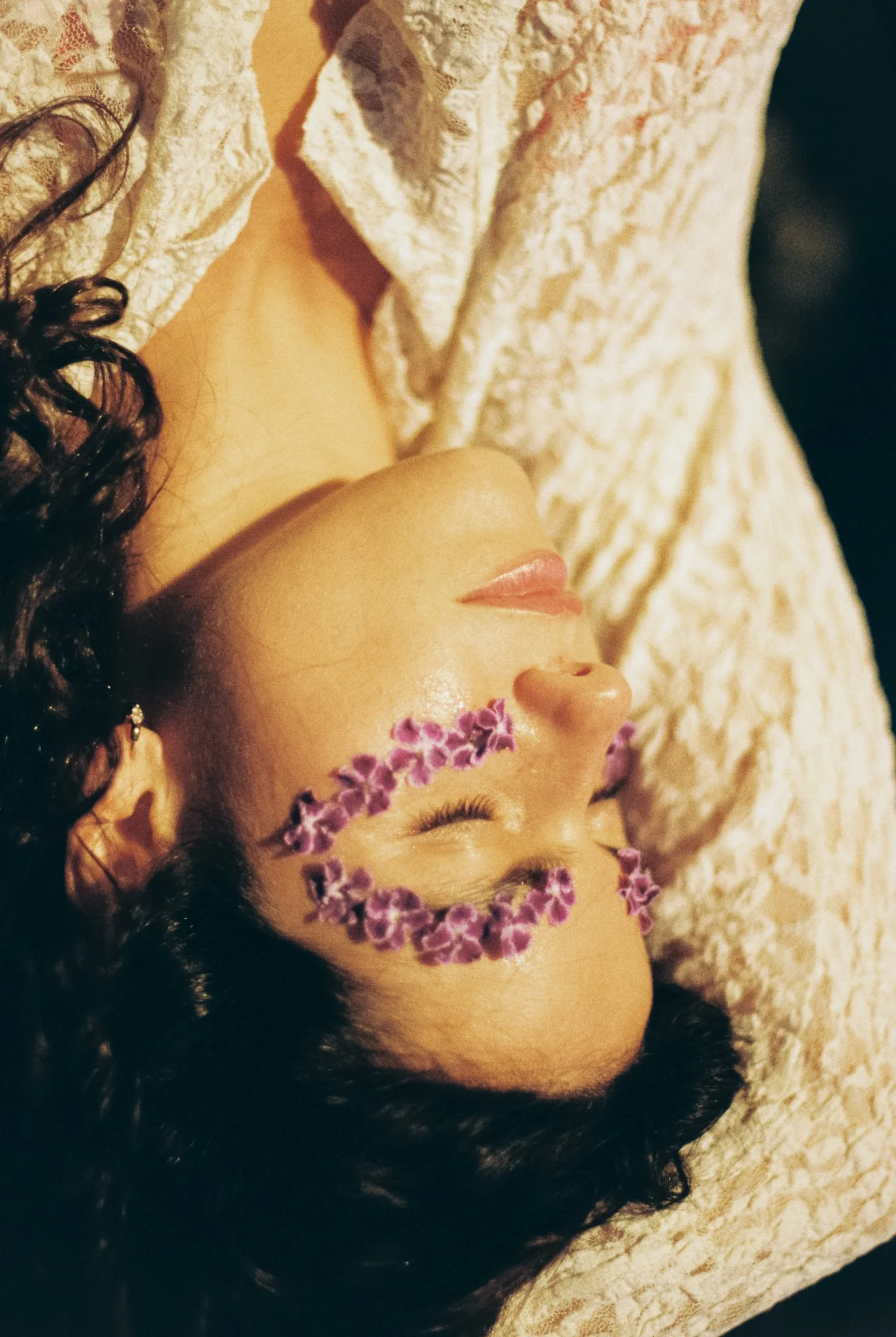 A woman with dark, curly hair lying on her side with her eyes closed. She has a crown made of small purple flowers on her forehead, and she is wearing a light-colored, textured fabric garment.