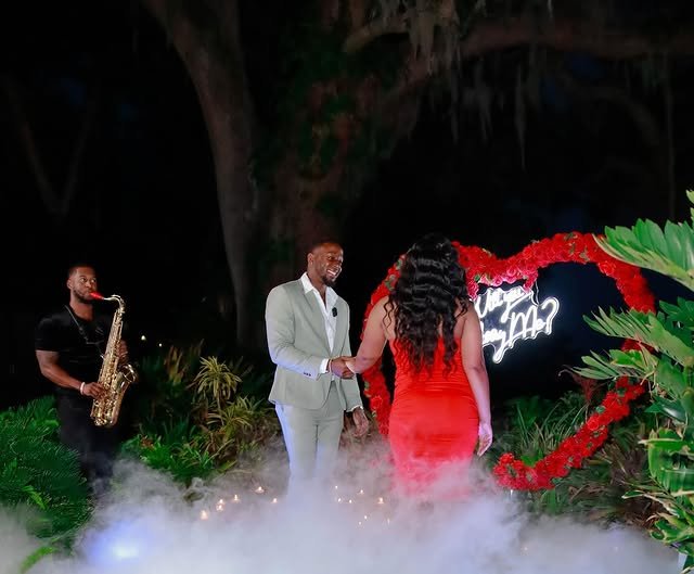 A couple holding hands and exchanging rings at an outdoor nighttime wedding ceremony decorated with a large red heart and neon sign, with a musician playing saxophone nearby, surrounded by lush greenery and mist.