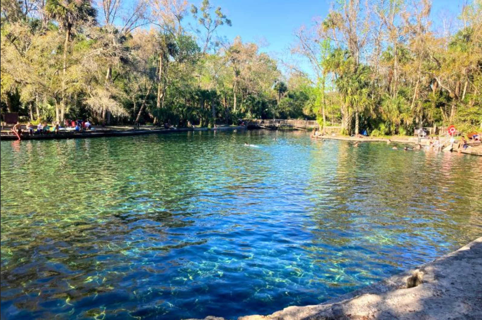 A serene river scene with clear blue water, surrounded by lush green trees and a few people lounging by the riverbank on either side.
