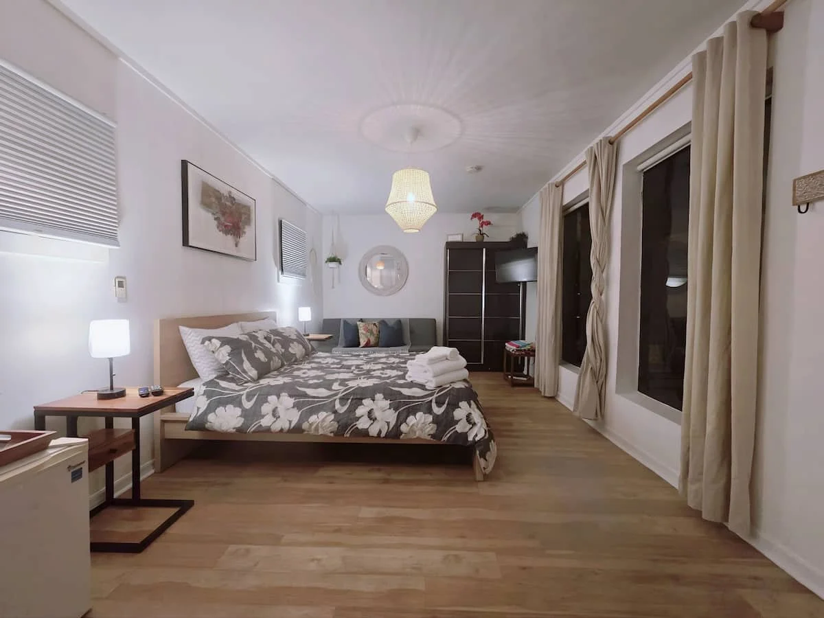 A cozy hotel room with a bed, bedside lamps, framed wall art, a sitting area, and large windows with curtains.