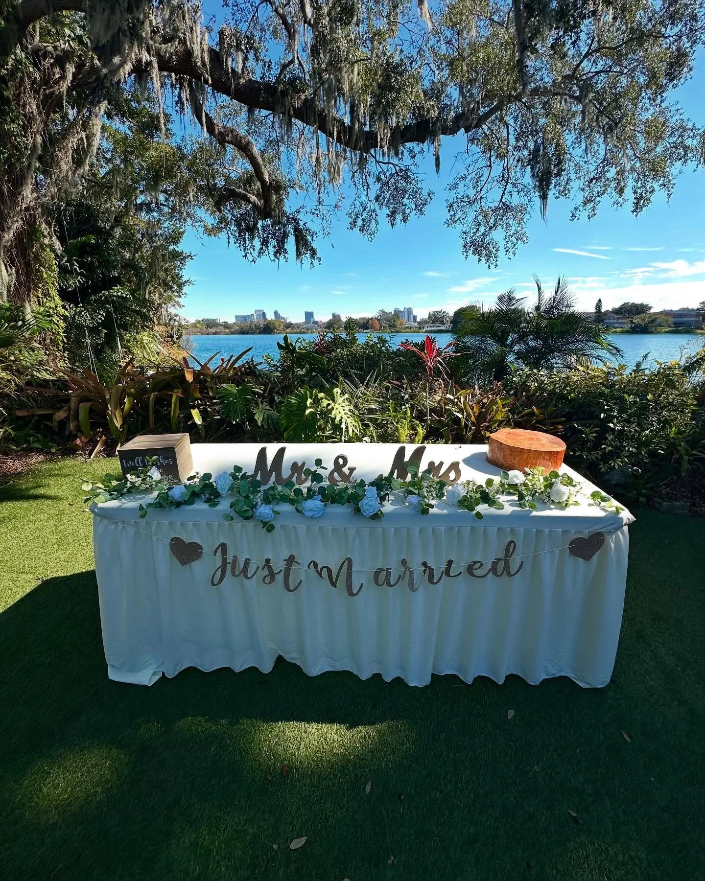 Wedding reception table with a white cloth, decorated with flowers, sign reading "Mr. & Mrs." and "Just Married", with a lakeside view, trees, and buildings in the background under a blue sky.