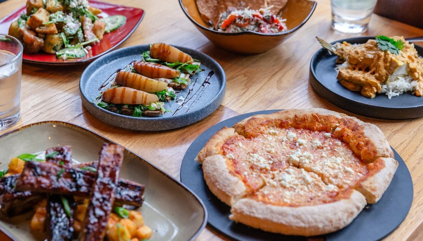 Various dishes on a wooden table including a pepperoni pizza, a salad, and different plates of Asian food.