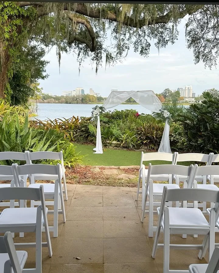 Wedding ceremony site set up outdoors by a river, with white chairs arranged for guests, a white canopy arch decorated with flowers, and lush greenery surrounding the area.