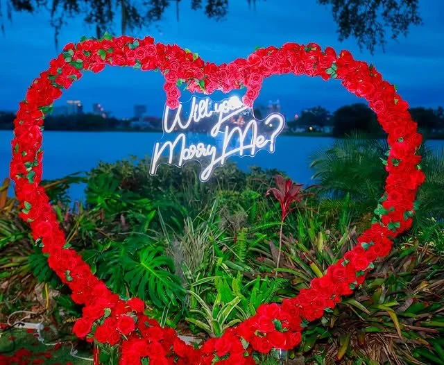 Heart-shaped floral arrangement of red roses with a neon sign inside that reads 'Will you marry me?' near a body of water and greenery.