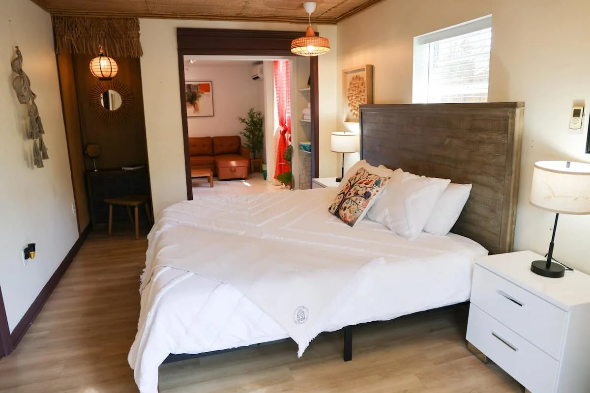 A bedroom with a large bed that has white bedding and decorative pillows. There are nightstands with lamps on either side. The room has a window with blinds, wooden floors, and a wooden headboard. In the background, a living area with a brown couch, 