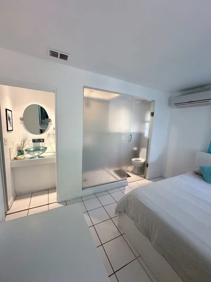 A bedroom with a white bed, a small blue pillow, and an air conditioning unit on the wall. There's a separate small bathroom with a frosted glass sliding door and a toilet visible inside. Adjacent to the bathroom is a vanity area with a round mirror,