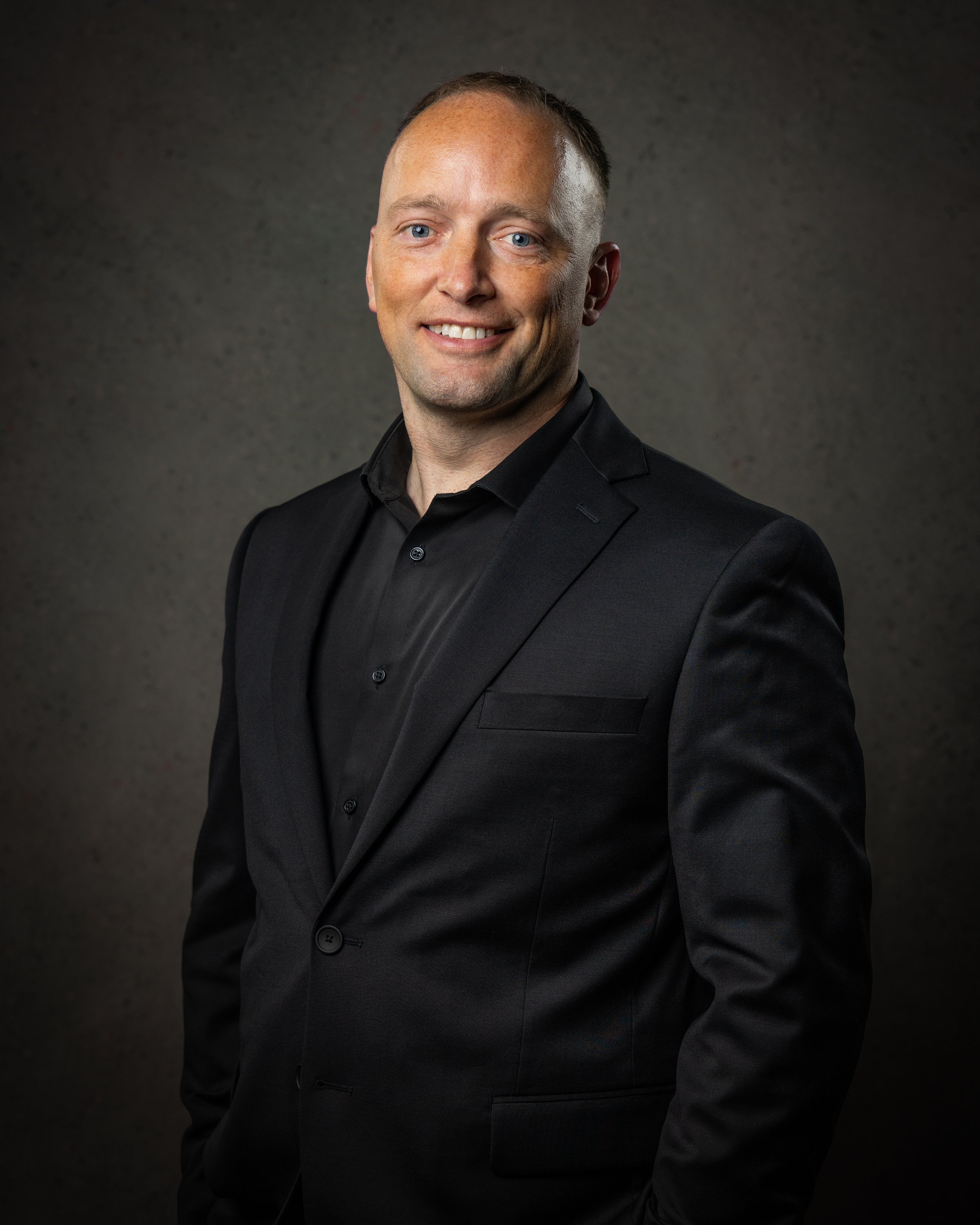Smiling man in black polo shirt with company logo, standing against dark background, arms crossed.